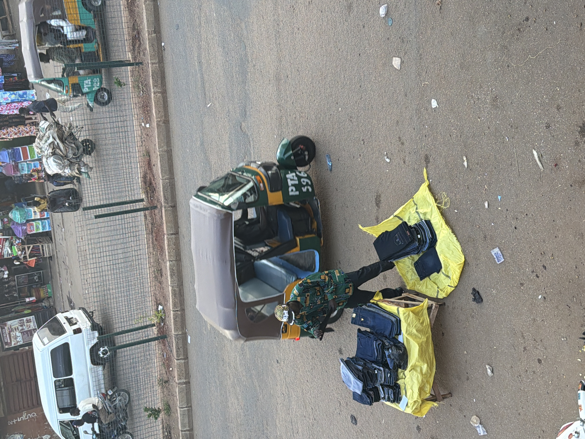 The image depicts an outdoor market scene in Bukuru, Nigeria. The primary subjects are a man dressed in a green and yellow patterned shirt and a patterned cap, standing next to an auto-rickshaw. He appears to be a vendor, as stacks of blue jeans are displayed on a yellow tarp in front of him. The auto-rickshaw, a common mode of transport in the region, is green and yellow and has "PTA 596" visible on its side.
Behind the vendor and auto-rickshaw, a white van is parked. To the left of the van, there are market stalls with various items, including what appear to be textiles and packaged goods, suggesting a bustling commercial area. A metal fence separates the market stalls from the road. The ground is paved with asphalt, and there are scattered pieces of debris.
The lighting suggests it is daytime, possibly late morning or afternoon, with no strong shadows indicating a specific time. The weather appears to be clear. The overall impression is of a vibrant street market where goods are being sold. There is no indication of a specific event, but the scene captures a typical day in this Nigerian location.