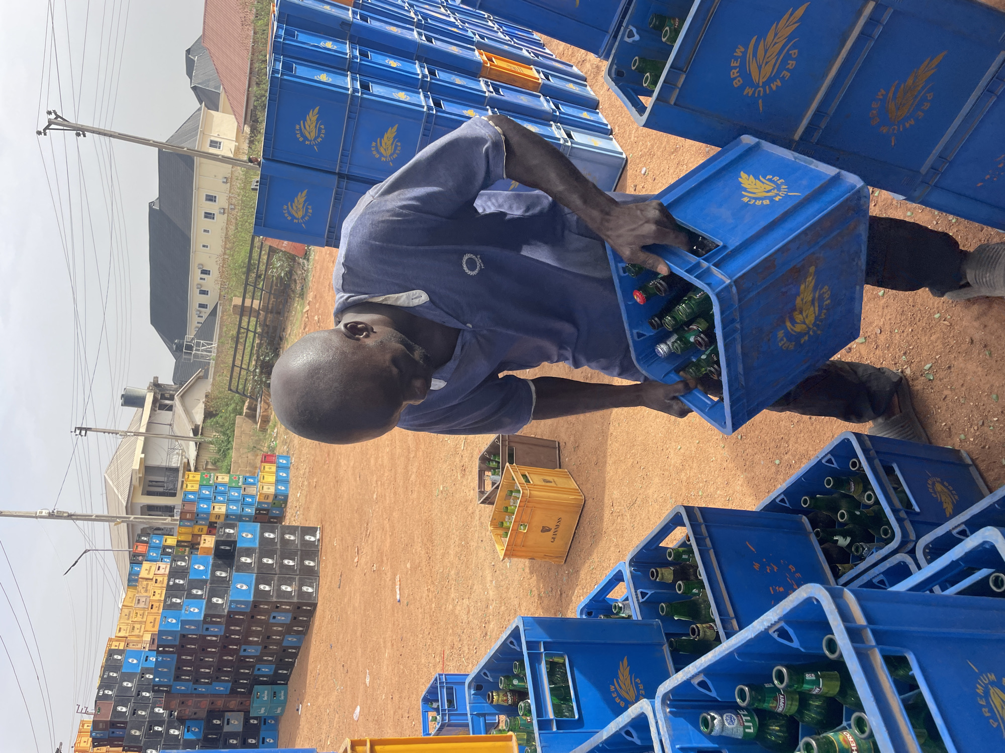 The image shows an individual in Gura Top, Nigeria, lifting a blue crate filled with green glass bottles. The person is wearing a blue short-sleeved shirt and gray pants. The ground is reddish-brown dirt. Surrounding the individual are multiple blue crates stacked in various configurations, many containing green glass bottles. Some yellow and brown crates are also visible. In the background, there's a building with a dark gray roof and beige walls, along with utility poles and power lines. The sky is overcast.