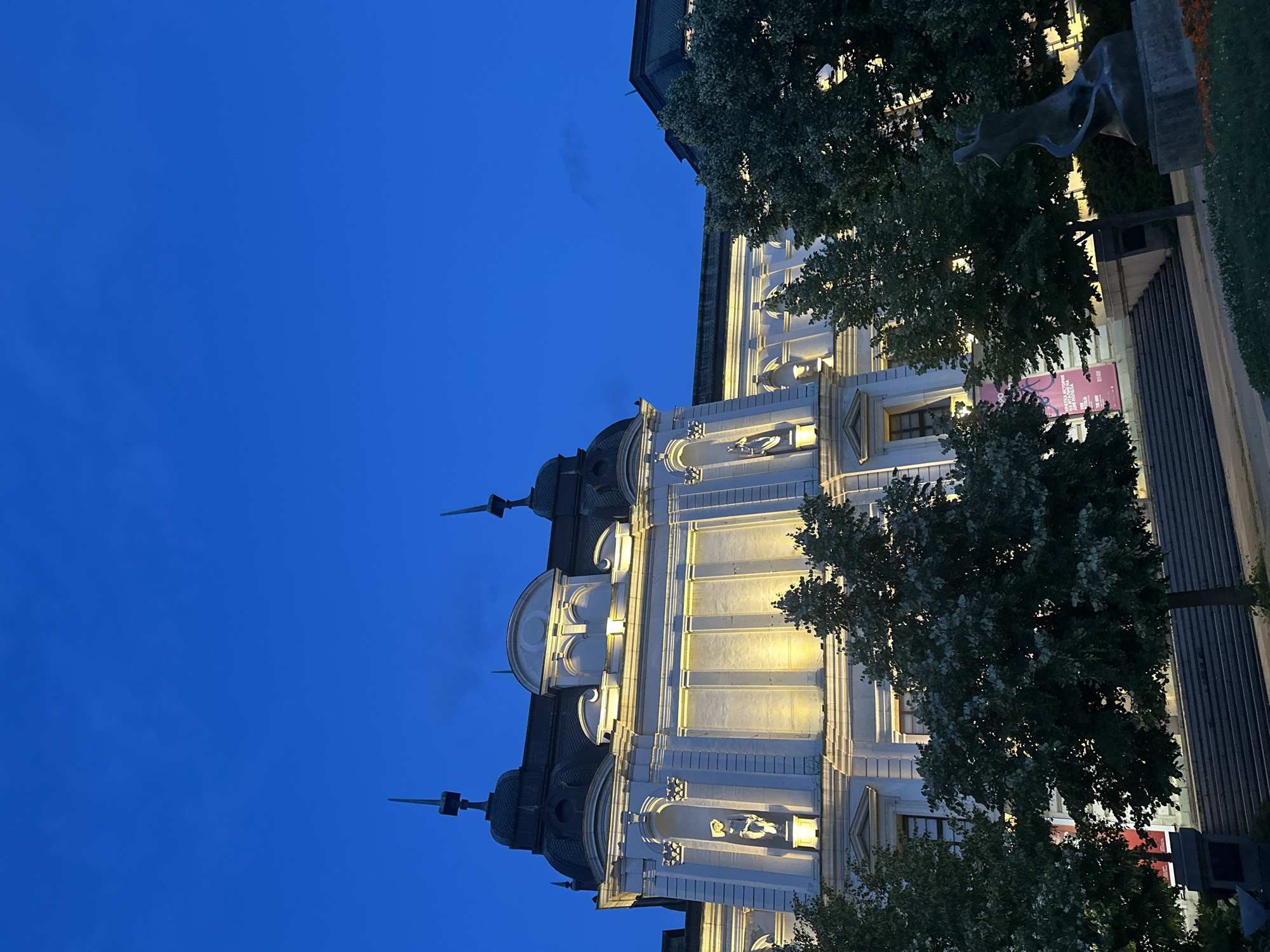 An exterior, low-angle, eye-level, full shot captures a large, classical-style building illuminated by artificial lights against a deep blue twilight sky. The building, painted white, features ornate architectural details, including sculptures in niches, arched windows, and a domed roof with spires. Trees with dense foliage are positioned in front of the building and to the right, partially obscuring the lower facade and steps leading up to it. In the foreground, to the right, a dark bronze statue of a figure in a dynamic pose stands on a pedestal. A pink poster is visible on the building to the left of the trees. The ground in front of the building is a paved area with some grass and flowerbeds. The overall scene depicts an urban architectural landmark at dusk or early night.