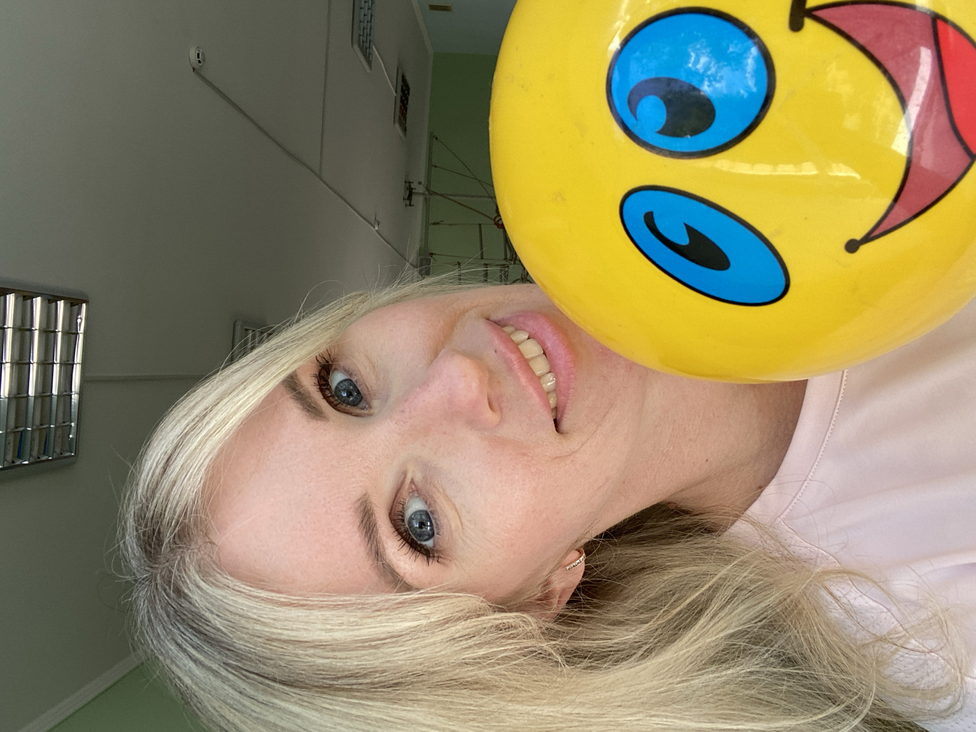 A woman with blonde hair and blue eyes is taking a selfie. She is holding a yellow balloon with a smiley face on it, which is partially obscuring her face. She is wearing a light pink t-shirt. The background appears to be an indoor space with a ceiling light fixture and some equipment in the distance. The image is taken from a close-up perspective, focusing on the woman and the balloon.