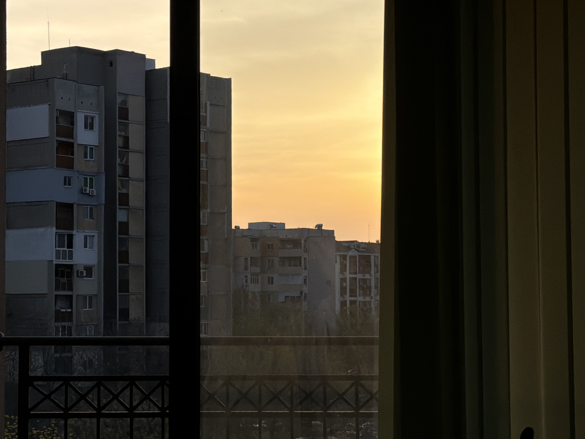 View through a window in Plovdiv, Bulgaria. The frame partially obscures a view of several concrete apartment buildings. The sky is a pale yellow, suggesting either sunrise or sunset. A balcony railing is visible in the lower portion of the image. Curtains are visible on the right side of the window frame. The buildings appear typical of Soviet-era construction.