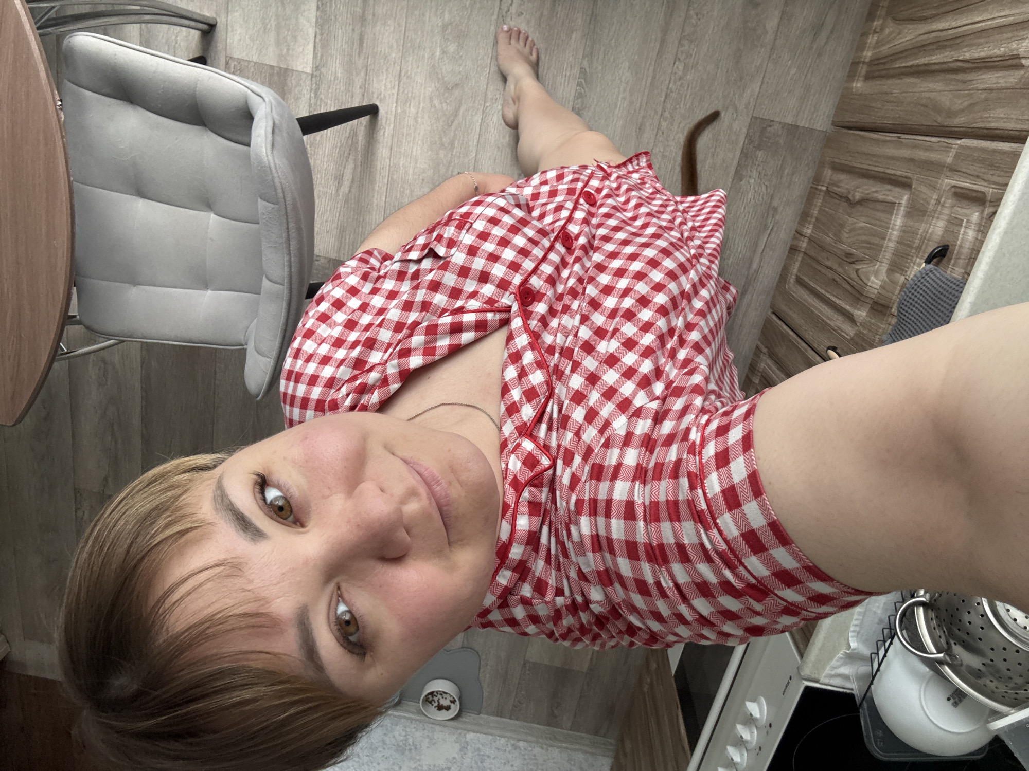 A woman is lying on the floor of an apartment in Stary Oskol, Russia. She is wearing a red and white checkered pajama set and has short, blonde hair. Her face is turned towards the camera, and she is smiling slightly. In the background, there is a gray chair, a round table, and a kitchen counter with a stove and dishes. The floor is made of light-colored wood-patterned laminate.