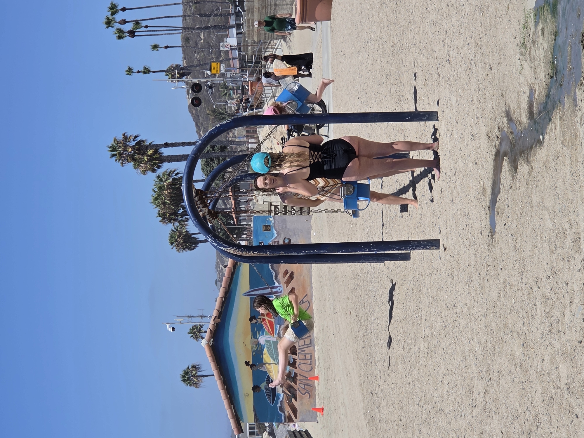 The image depicts a sunny day at San Clemente State Beach in California. In the foreground, a woman in a black swimsuit is swinging on a swing set. She has blonde hair and is wearing a turquoise helmet. Behind her, a young girl in a green shirt and shorts is also on a swing, her feet dangling as she moves. The swing set has a blue frame.
In the background, there is a mural painted on a wall that appears to depict surfers and waves. Palm trees are visible, and a bright blue sky suggests fair weather. There are also railway tracks with signals, indicating the proximity of a train line. Further in the distance, a hillside with sparse vegetation can be seen. The ground is covered in sand and gravel.
The overall atmosphere seems relaxed and recreational, fitting for a beach environment. The lighting suggests it is midday. Text is visible on the mural, spelling out "SAN CLEMENTE," and a yellow sign with "RAILROAD" and "TRAIN" is also present.