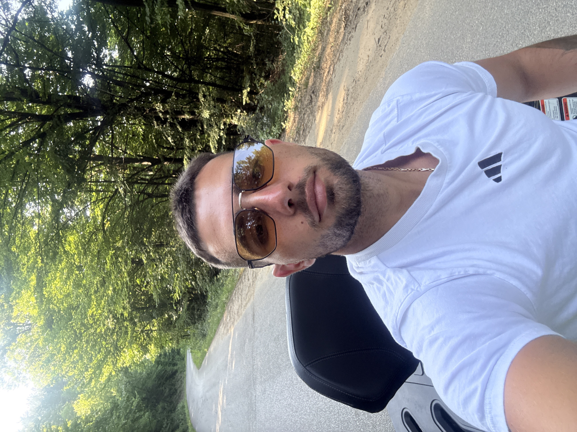 A man, with short dark hair and a beard, is taking a selfie while sitting on a motorcycle. He is wearing a white t-shirt with the Adidas logo on the left side of the chest and a gold chain necklace. He has on sunglasses with brown lenses. The motorcycle seat is black and appears to be made of leather.

The man is outdoors, on a paved road that winds through a forest. The trees are lush and green, with sunlight filtering through the leaves, suggesting it is daytime, likely late morning or early afternoon. The road is bordered by a dirt path on the right. The overall impression is one of a scenic ride through a natural landscape, possibly in Orlovtsi, Bulgaria, given the location context. The lighting and the man's relaxed posture suggest a pleasant day for a motorcycle excursion. There is no visible text in the image.