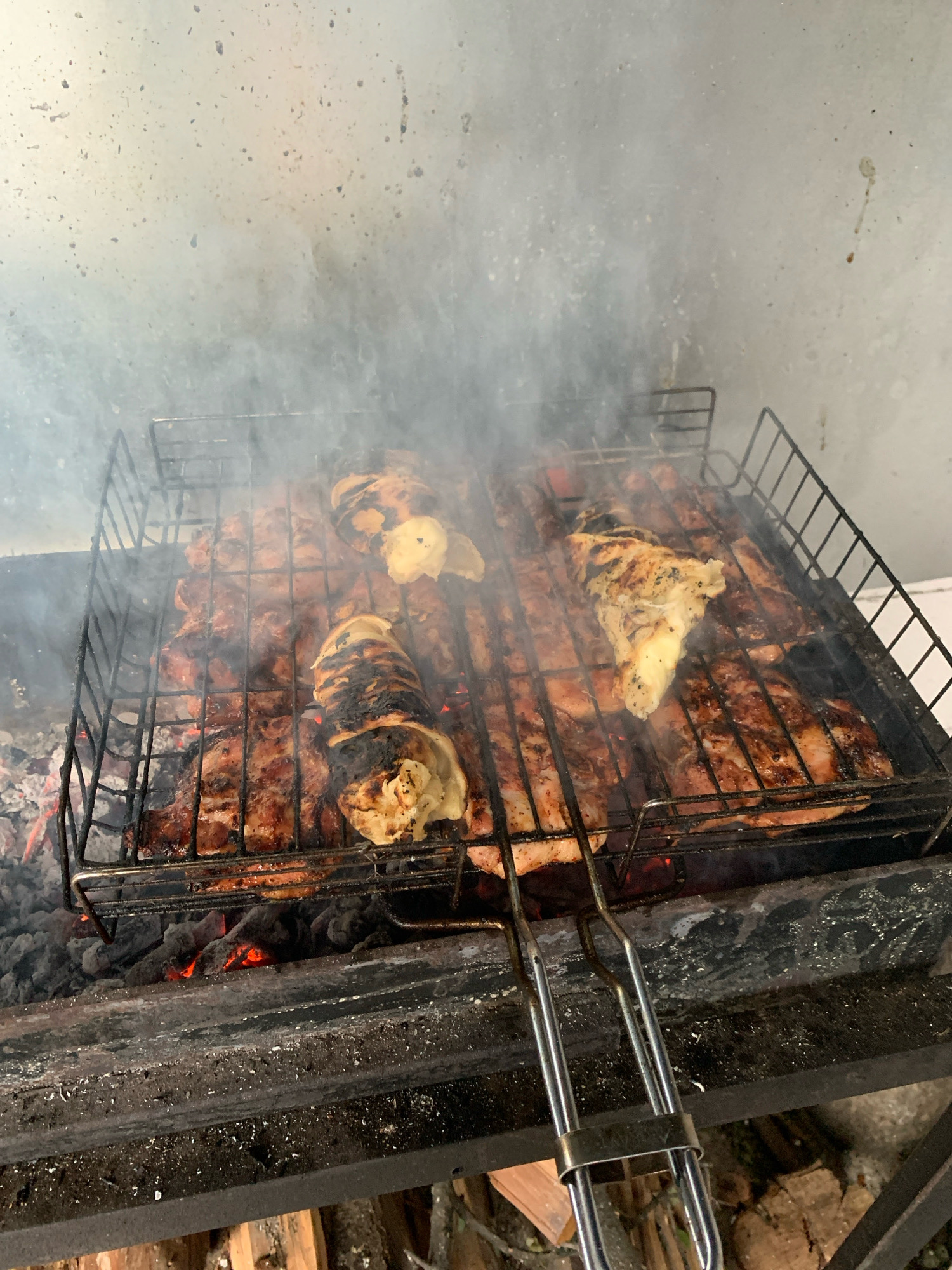 This image depicts an outdoor cooking scene, specifically a barbecue with meat grilling over hot coals. The primary subjects are numerous pieces of raw and partially cooked meat arranged on a metal grill basket. The meat appears to be various cuts, possibly pork and chicken. Some pieces show charring and browning, indicating they are in the process of being cooked. Smoke is rising from the grill, obscuring some of the details of the meat and the background.
The grill itself is a standard wire mesh basket with handles, placed over a bed of glowing red coals. The coals are visible at the bottom of the image, emitting heat and smoke. The grill is situated within a metal barbecue pit or structure, which appears to be made of dark metal and shows signs of use, with some soot and residue. The background is a light-colored, textured wall, possibly concrete or a similar material, splattered with dark marks, likely from cooking grease or smoke.
The scene suggests a domestic or casual outdoor gathering. The presence of what appears to be stacked firewood in the lower right portion of the image further indicates an outdoor setting, likely in a backyard or garden. The overall impression is one of food preparation and outdoor leisure.
Without additional context or visual cues, it is not possible to definitively pinpoint the location within the city of Sumy, Ukraine. However, the style of the barbecue and the general appearance of the surroundings are consistent with a typical outdoor residential or recreational area found in many Ukrainian cities.