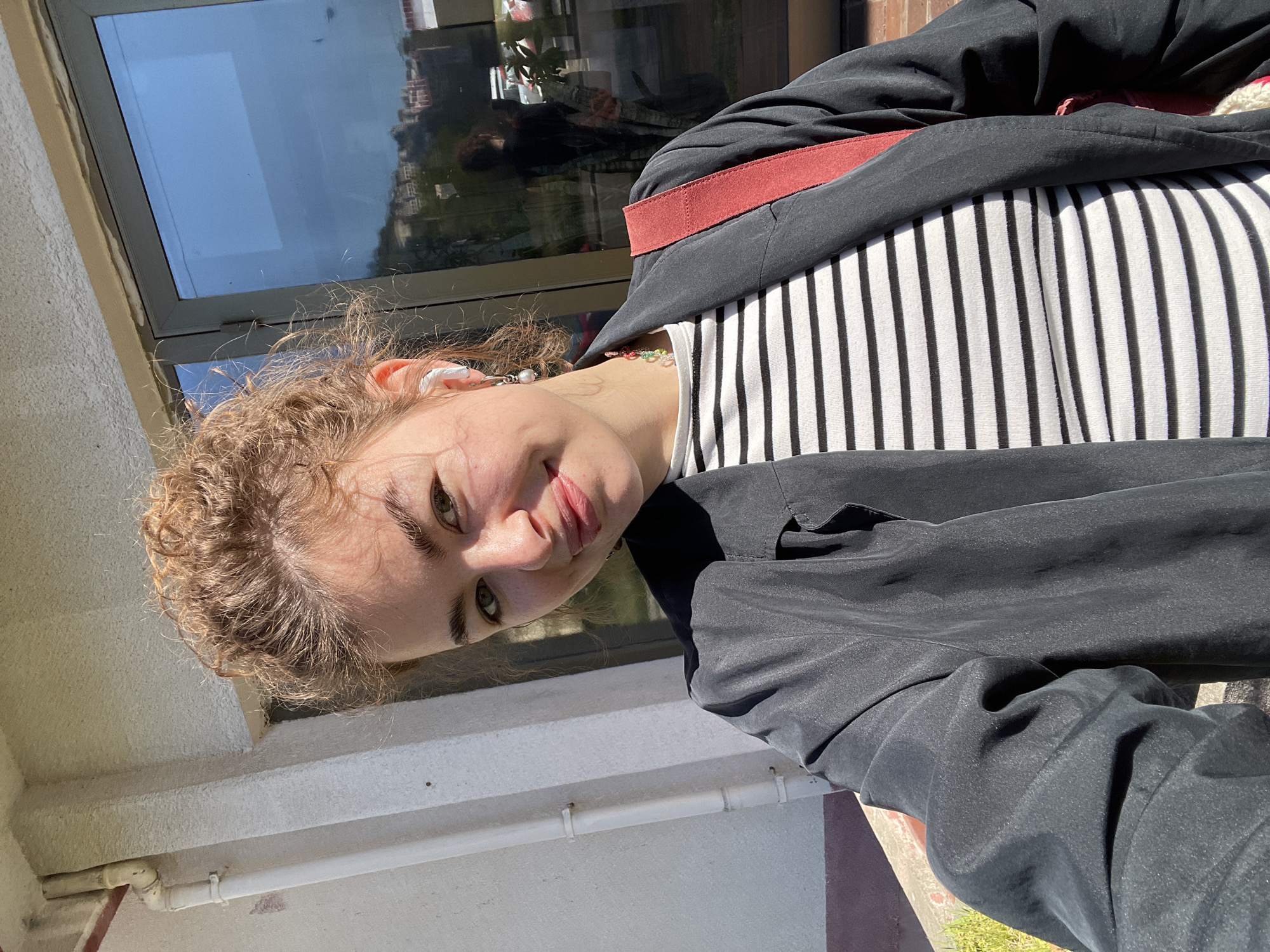 A young woman with curly hair is photographed outdoors, likely in Budva, Montenegro. She wears a black blazer over a black and white striped shirt. A red strap is visible across her shoulder. The background features a window reflecting buildings and sky. The lower part of the frame shows a concrete building exterior and hints of green vegetation. The image is slightly tilted counter-clockwise.