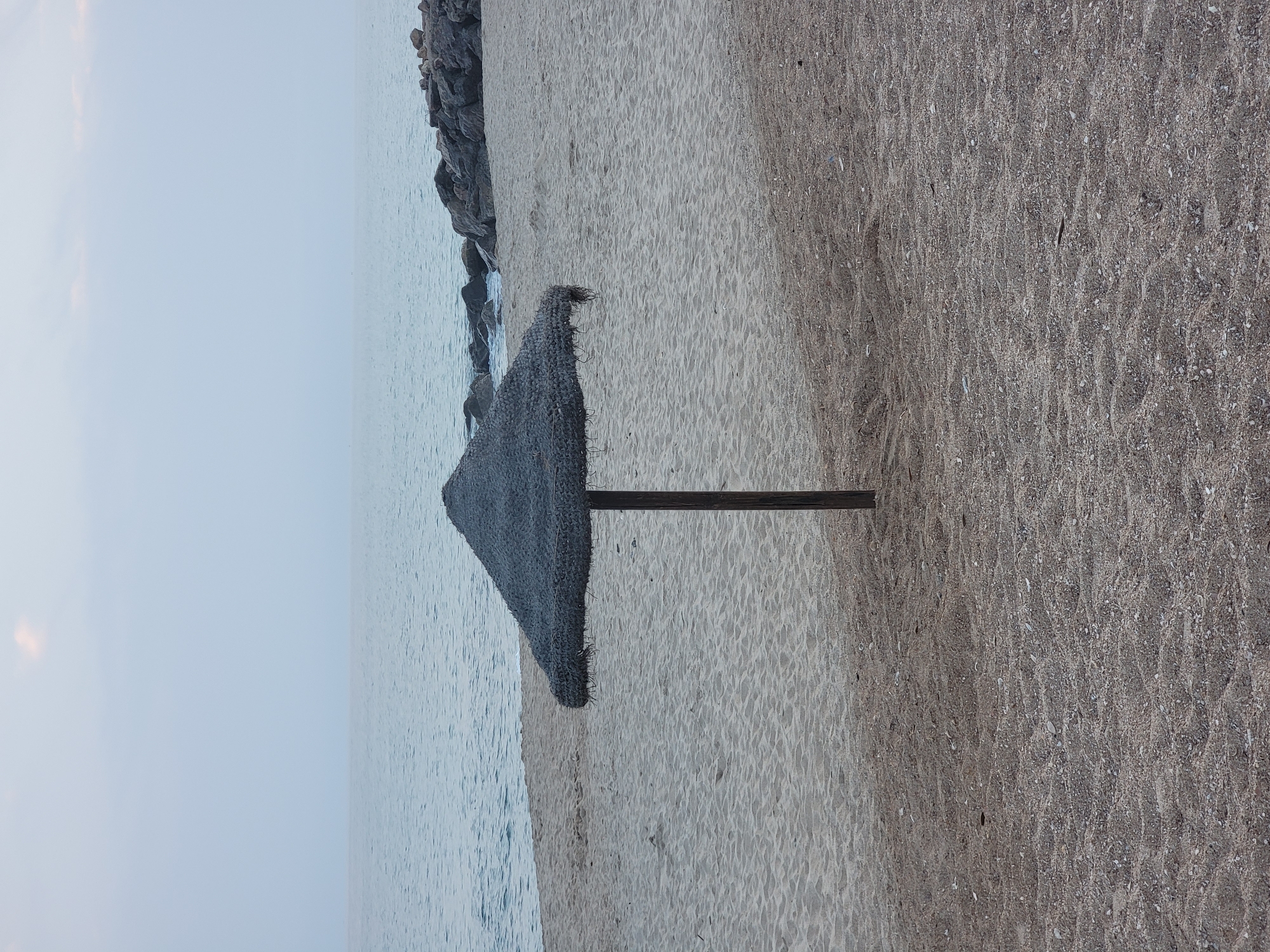 A thatched umbrella stands on a sandy beach in Quarteira, Portugal, with the calm sea stretching to the horizon under a pale sky. The umbrella, made of dark thatch, is supported by a single wooden pole and casts a shadow on the sand. The beach is sparsely populated, with visible rocks forming a breakwater in the distance. The sand is a light brown color, with a coarser texture in the foreground and finer grains closer to the water. The sky is overcast with a hint of soft pink, suggesting either dawn or dusk.