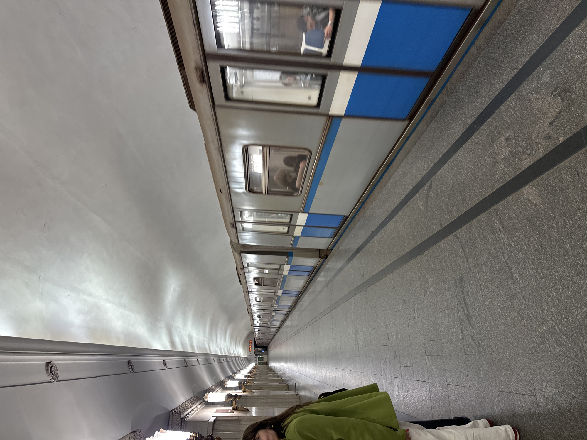 The image captures a long, deep underground subway station platform, characteristic of the Moscow Metro. A grey subway train, adorned with a prominent blue stripe, is depicted in motion, likely arriving or departing, with its movement evident through the blur of its windows and visible passengers.
The station platform extends far into the distance, defined by a high, vaulted white ceiling and a textured grey tiled floor featuring dark parallel lines that delineate the track area. Along the left wall of the tunnel, regularly spaced ornate lighting fixtures, possibly made of bronze, illuminate the pathway. The perspective emphasizes the impressive depth and length of the tunnel, leading to a distant, brightly lit section where a sign with indistinct red lettering can be faintly seen.
In the foreground, partially visible on the lower right side of the platform, is a person wearing a vibrant green jacket and dark hair. Only their upper torso and head are visible, suggesting they are standing or sitting on the platform, possibly observing the train or the station. The scene conveys the bustling atmosphere of an urban transportation hub. Due to the underground setting, no specific time of day or weather conditions are discernible.