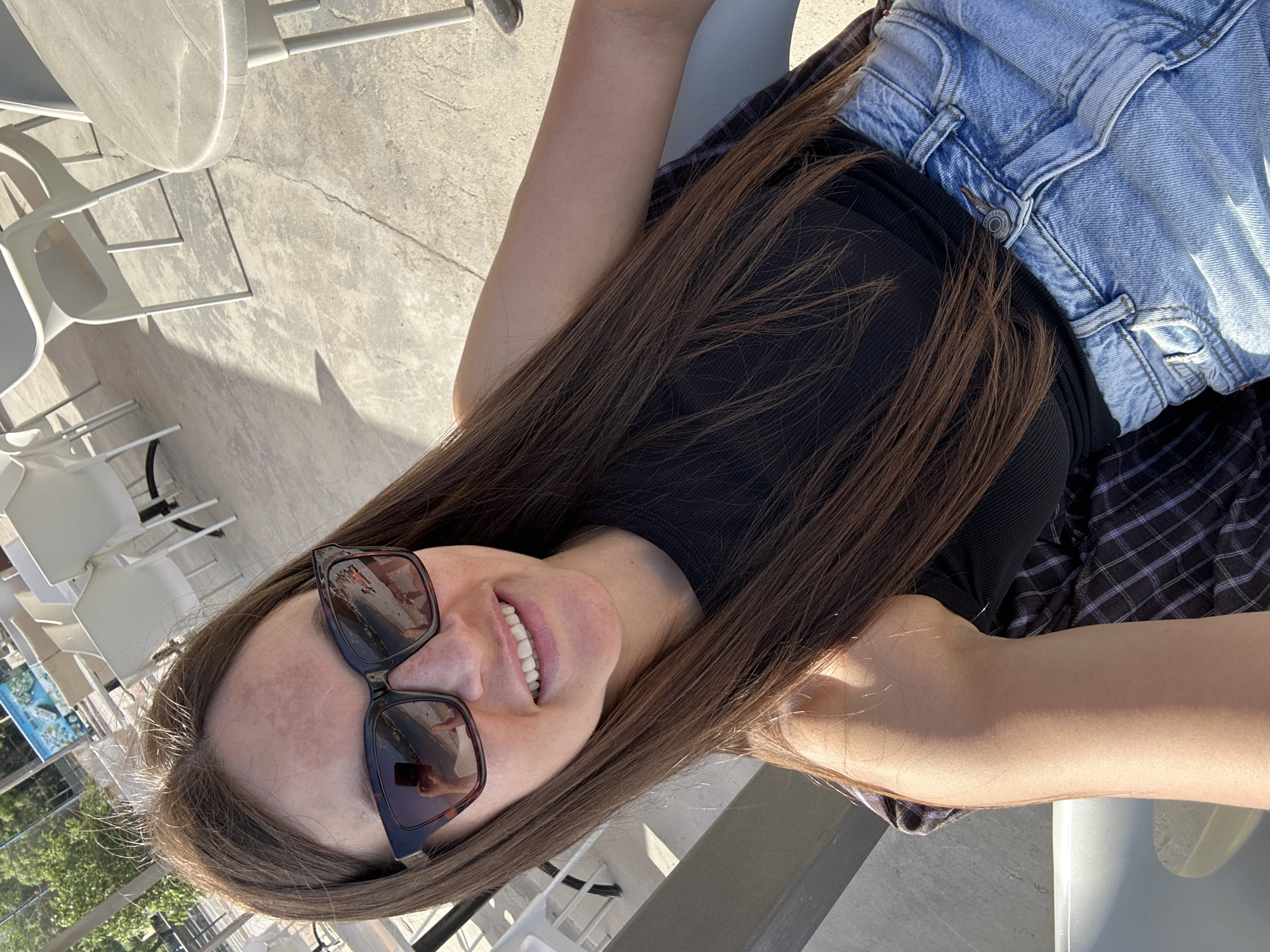 A young woman is photographed from a close, high-angle perspective. She is wearing blue jeans, a black top, and sunglasses. She is smiling, and her long brown hair is spread around her. The background shows a terrace or patio with white chairs and tables. A sign with a picture is partly visible behind her, suggesting a location with a view. The photo likely taken in Budva, Montenegro, judging by the outdoor setting and potential coastal location.