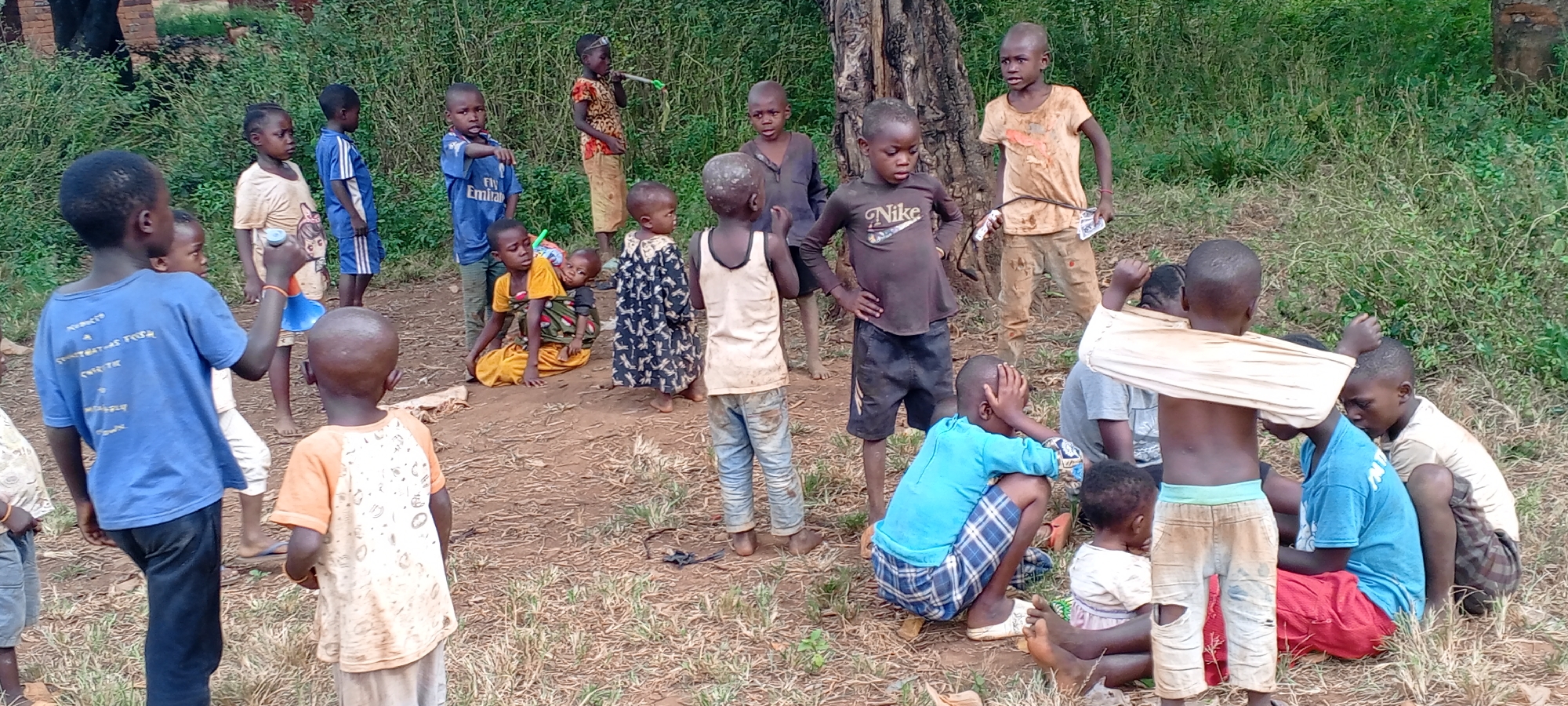 In Ngalula, Democratic Republic of the Congo, a large group of children are gathered outdoors. The children are barefoot and wearing an assortment of worn clothing, including t-shirts with logos, patterned dresses, and jeans. Some children are standing, while others are sitting or crouching on the bare earth. A few of the children are interacting with each other. The background consists of dense green vegetation and trees, with hints of brick structures visible in the distance. The ground appears to be a mix of dirt and sparse grass.