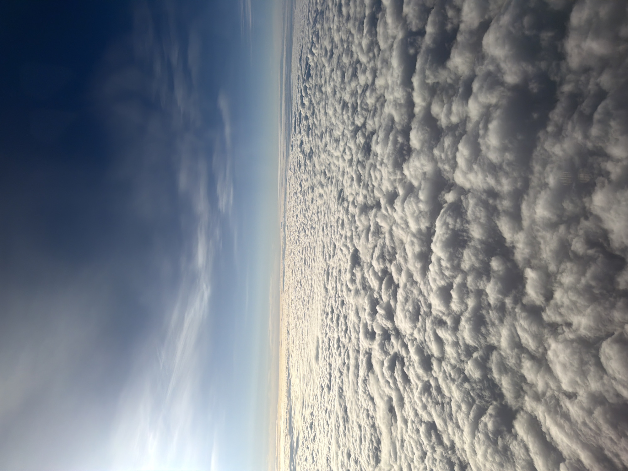 An aerial view shows a vast expanse of white, puffy clouds filling the lower half of the frame. These clouds appear to form a dense, undulating layer, obscuring whatever lies beneath. The upper half of the image is dominated by a gradient of blue sky, transitioning from a lighter, hazy hue near the horizon to a deep, rich blue at the top. wispy, thin clouds are visible in the upper sky. The sun is visible in the top left corner, casting a bright light that illuminates the cloud layer, creating highlights and shadows that give the clouds a three-dimensional appearance. The horizon line is clearly defined, separating the cloud layer from the sky. There are no people, structures, or any indication of the city of Gardermoen, Norway visible in this image; it is a purely atmospheric scene.