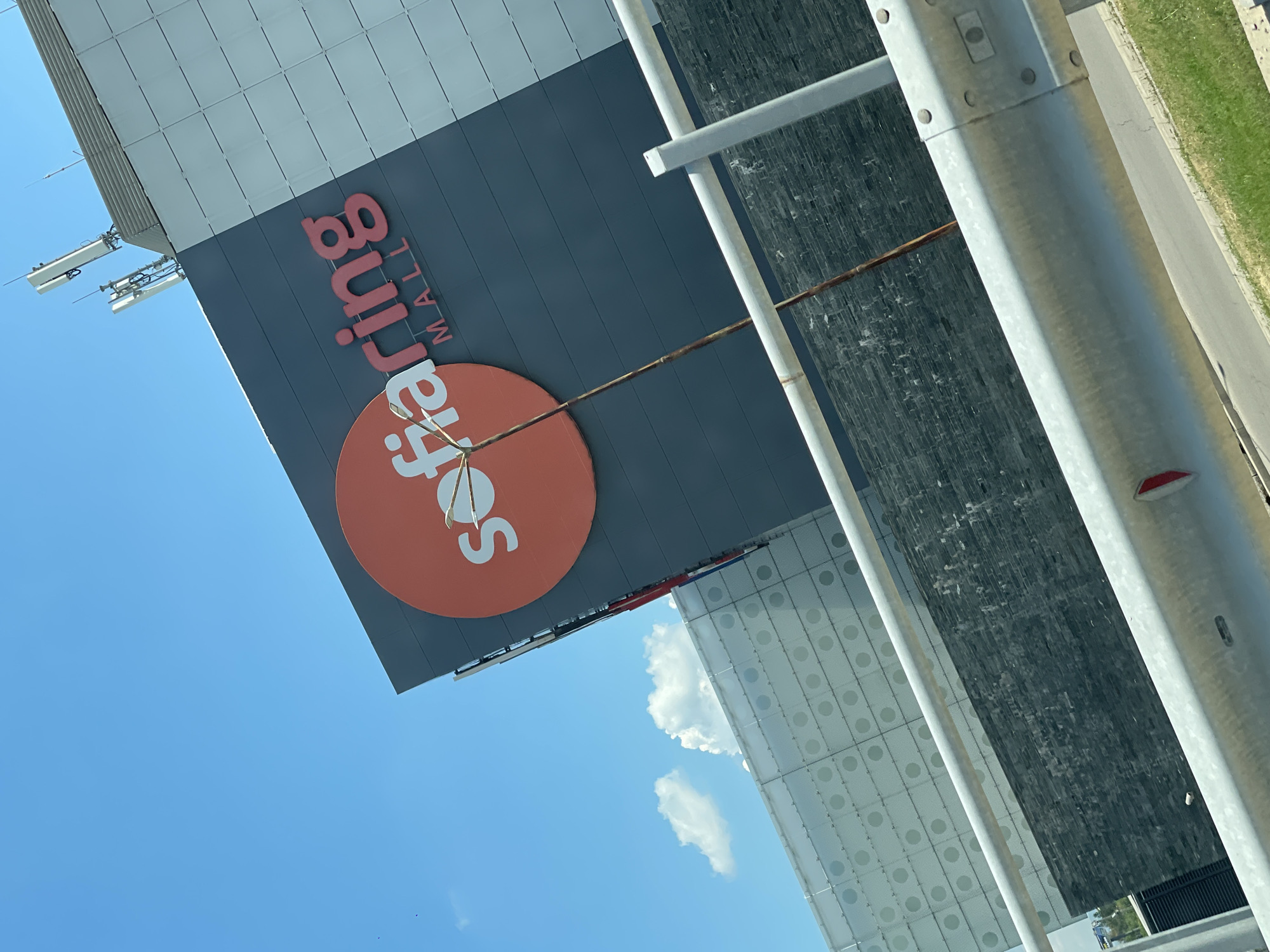 The image shows the Sofiaring Mall, a commercial building located in Sofia, Bulgaria. The mall features a prominent sign with the name "Sofiaring MALL" in red lettering against a dark grey facade. The building's exterior is characterized by a large orange circular logo with white text. The upper part of the building is clad in light-colored panels, with telecommunication antennas visible on the rooftop. In the foreground, a metal guardrail separates the viewer from the building. The sky is clear blue with a few scattered clouds. The scene is captured on a sunny day.