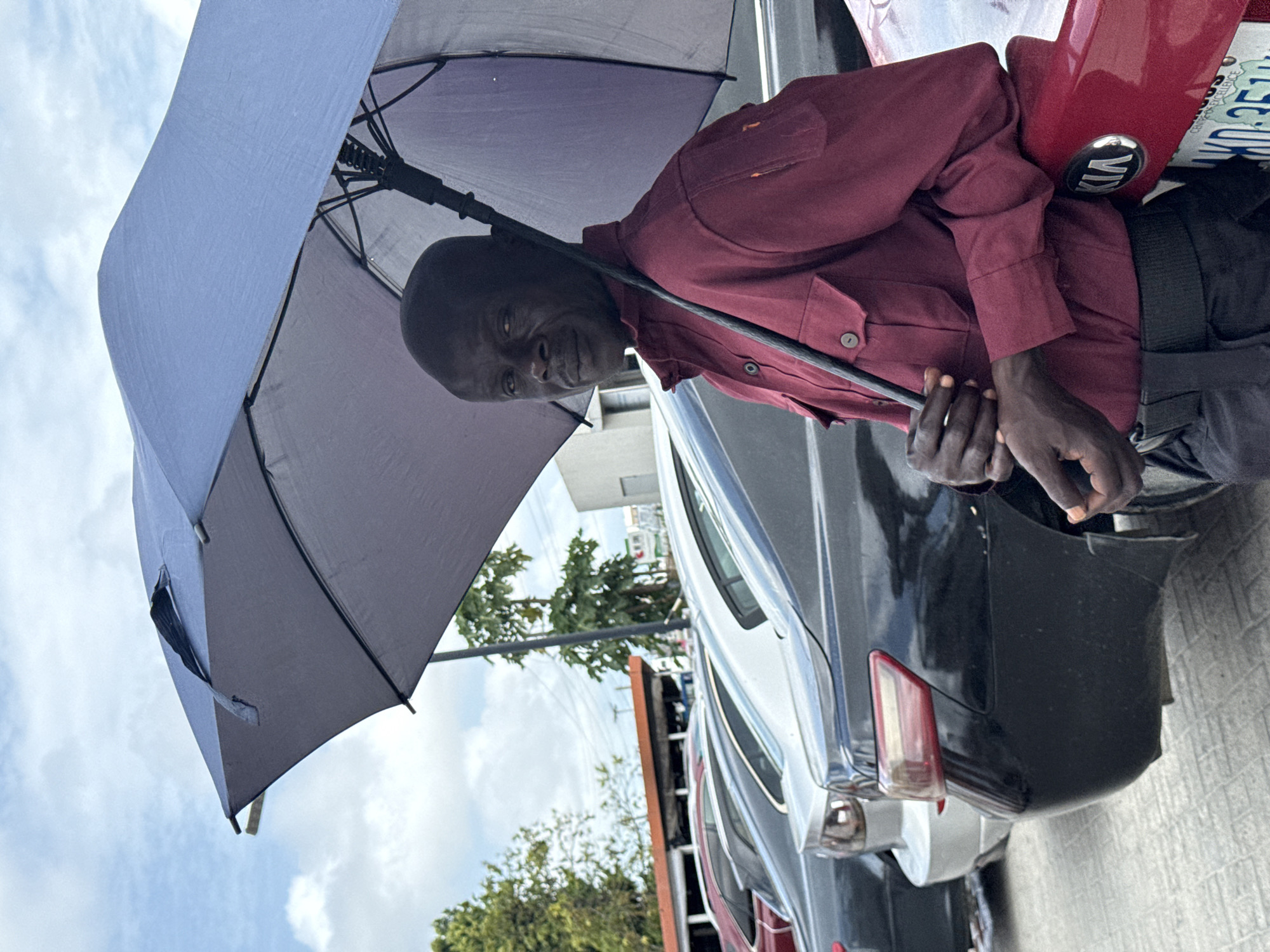 A medium shot captures a man holding an umbrella in Ado, Nigeria. He is wearing a maroon button-down shirt. The man stands beside a row of parked cars; the closest one is a black KIA with a red bumper, its license plate partially visible. The sky is visible behind the trees in the background. The ground is paved.