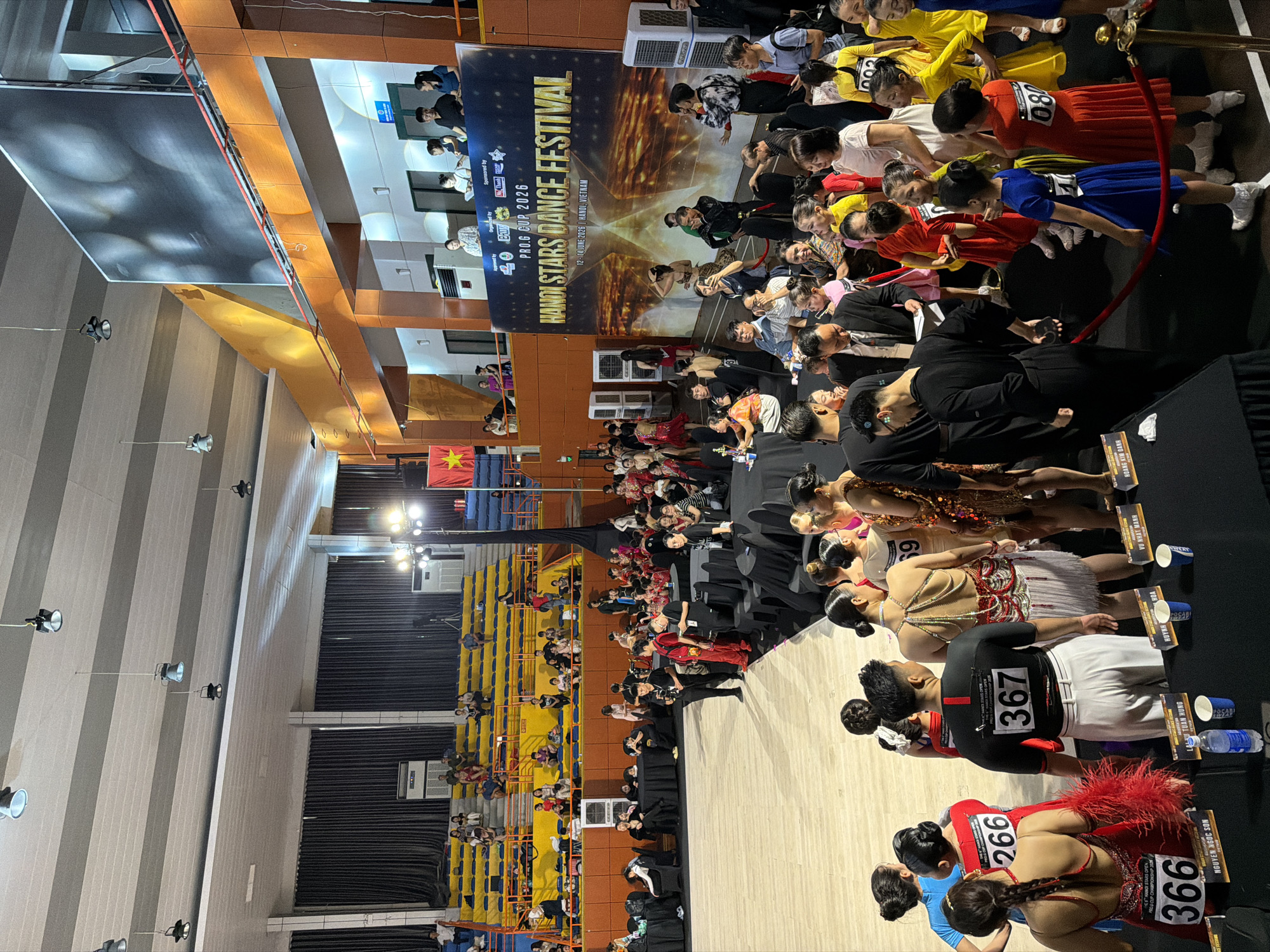 This image depicts a ballroom dance competition titled "HANOI STARS DANCE FESTIVAL" and "PRO.G CUP 2026" at the Cung Thể Thao Tổng Hợp Hà Nội (Hanoi Sports Palace). The scene is indoors, with a large digital screen in the background displaying event information and imagery. Rows of spectator seating are visible, some occupied by people. The foreground features a dance floor area where participants, many in elaborate costumes, are gathered. Several individuals appear to be competing or preparing to compete, with numbers displayed on their attire (e.g., "367," "266," "366," "080"). Other people, including organizers or staff, are interacting with the participants. Tables are set up with drinks and event materials. The overall atmosphere suggests a formal and energetic event.