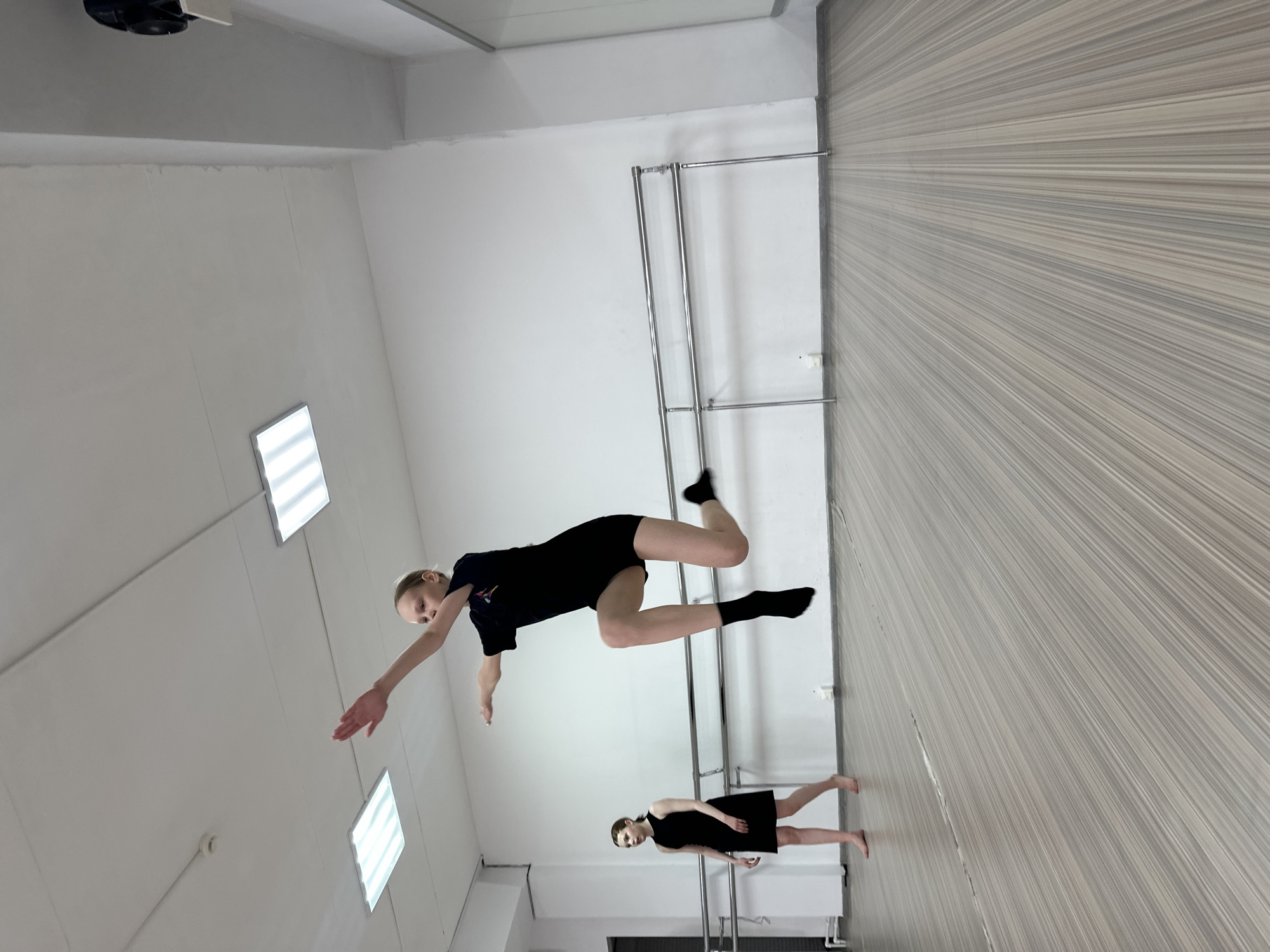 This image, taken in Votkinsk, Russia, captures two young dancers practicing in a brightly lit studio. The setting features white walls, a light-colored wooden or laminate floor with a distinct linear grain pattern, and rectangular panel lights illuminating the ceiling.
In the foreground, a blonde-haired girl, wearing a black t-shirt, black shorts, and black socks, is depicted mid-air, performing a dynamic dance movement or jump. Her body is horizontally oriented, with her left arm extended forward and legs bent. She maintains a focused expression, looking towards the viewer.
Further back in the studio, a second girl with dark hair, dressed in a black sleeveless dress, stands barefoot on the floor. She is positioned next to a silver ballet barre mounted against the white wall. A long, horizontal mirror is also visible on the wall behind her, reflecting portions of the room.
Other visible details include a ventilation fan or similar equipment in the upper left corner and a small white sensor or speaker attached to the ceiling. The studio appears to be a functional and clean space, well-suited for dance training. There is no discernible text in the image, and the artificial lighting means the time of day and weather are not apparent. Both dancers display concentration, reflecting their engagement in practice.