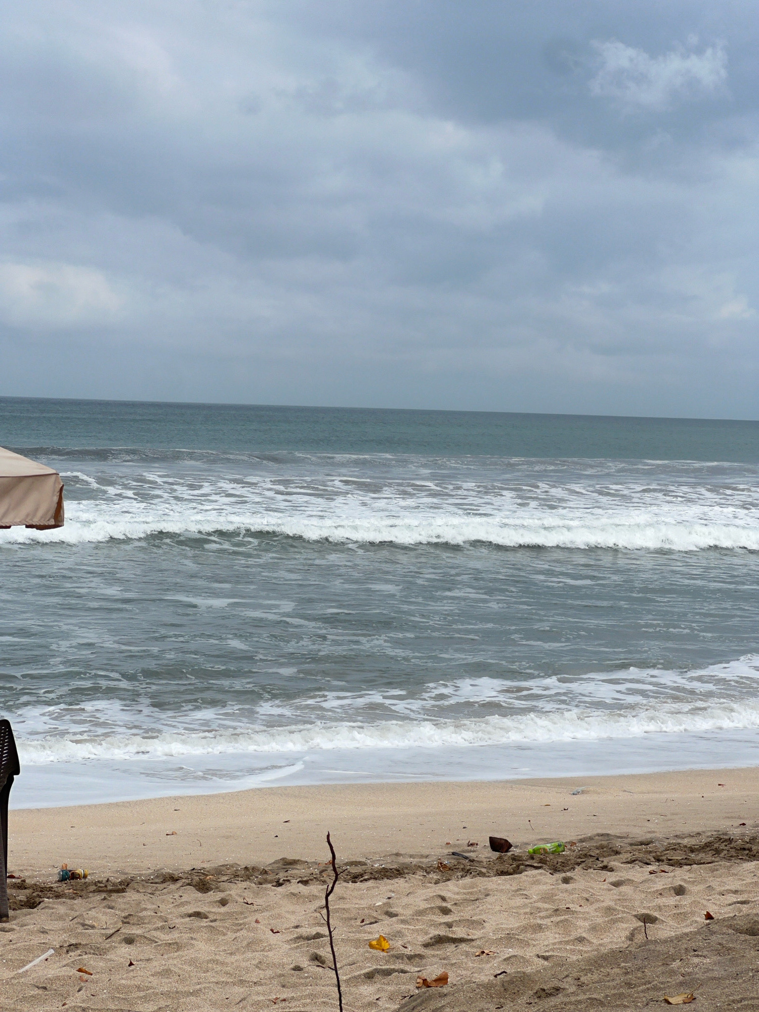 The image depicts a cloudy day at Kuta Beach, Indonesia. The foreground shows a sandy beach littered with debris, including plastic bottles and leaves. A dark, possibly metal, object is partially visible on the left, and a thin branch protrudes from the sand in the center. The middle ground features the ocean with white-capped waves rolling towards the shore. In the distance, the horizon line separates the sea from a heavily overcast sky. A section of a tan beach umbrella is visible in the upper left corner of the frame.