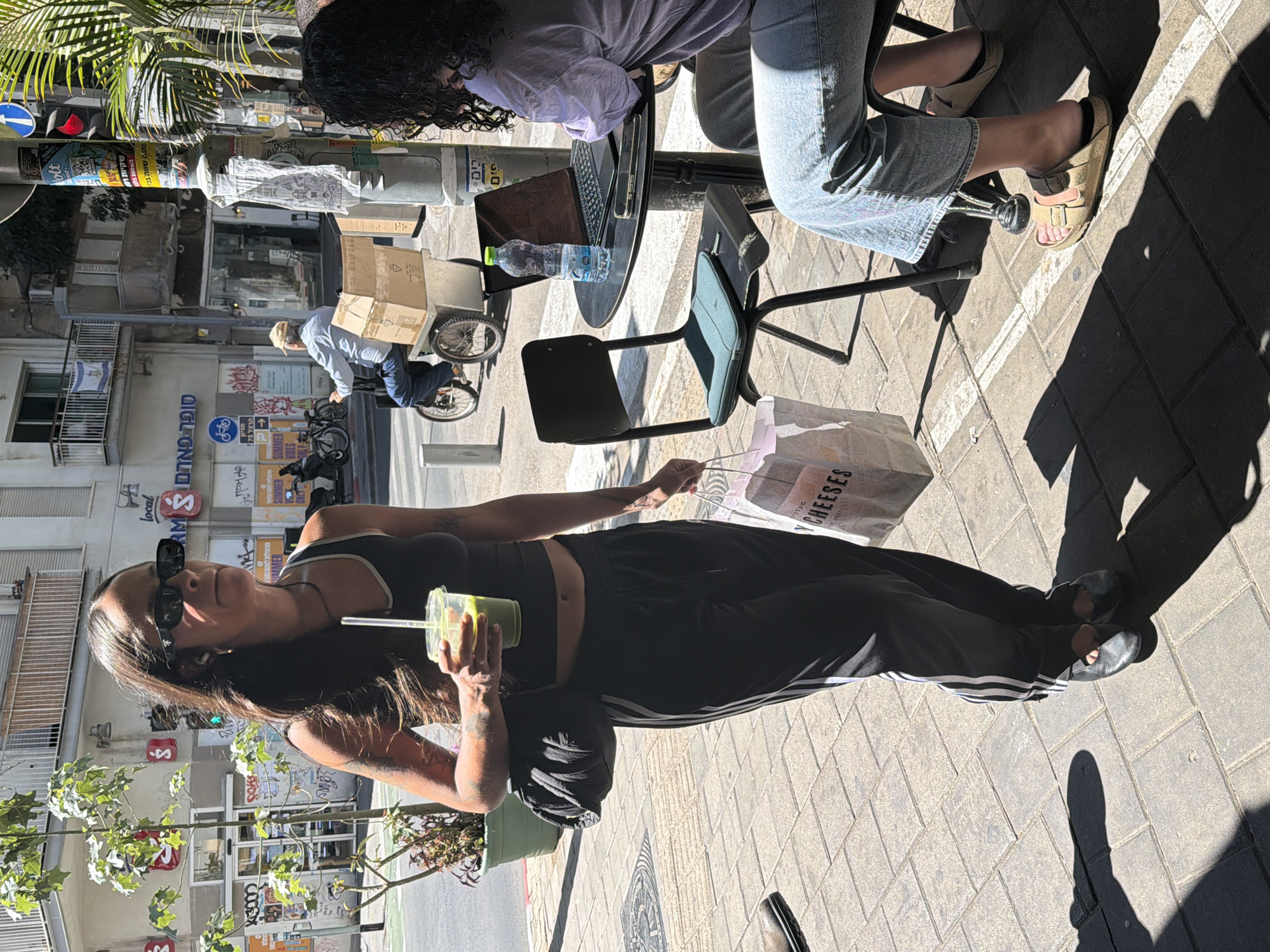Here is a detailed description of the image:
**General Impression:** The image captures a daytime scene on a street in what appears to be Tel Aviv, Israel. The setting appears to be a commercial or urban area.
**People:**
- The main subject is a woman standing in the foreground. She's wearing sunglasses, a black top, and black track pants with white stripes down the sides. She's holding a green drink (possibly a smoothie or juice) in her right hand and a shopping bag in her left.
- In the background, another person with curly hair is sitting at a table, seemingly working on a laptop. They're wearing a light-colored shirt and jeans.
- Further in the background, a person is riding what looks like a bicycle with a cart attached, carrying cardboard boxes.
**Objects:**
- A table and chairs are present, where the person with the laptop is seated. There's a bottle of water on the table.
- The woman is carrying a shopping bag with the word "CHEESES" printed on it.
- A green drink with a straw is held by the woman in the foreground.
- There are buildings, trees, street signs, and other urban elements visible.
**Actions and Interactions:**
- The woman in the foreground appears to be posing or simply standing and holding her drink.
- The person at the table seems to be working on a laptop.
- The person on the bicycle is transporting boxes.
**Environment:**
- The scene takes place on a paved street with brick patterns.
- There's a mix of sunlight and shadows.
- The buildings in the background have various signs and advertisements on them.
- There's a street sign indicating "Herzl 75", and a local ad for "SUMMER".
- There's a Super-Pharm sign.
**Location:** Based on the signs visible (Herzl 75, Super-Pharm), the location is likely in Tel Aviv, Israel. Specifically, it appears to be near or on Herzl Street (רצל).