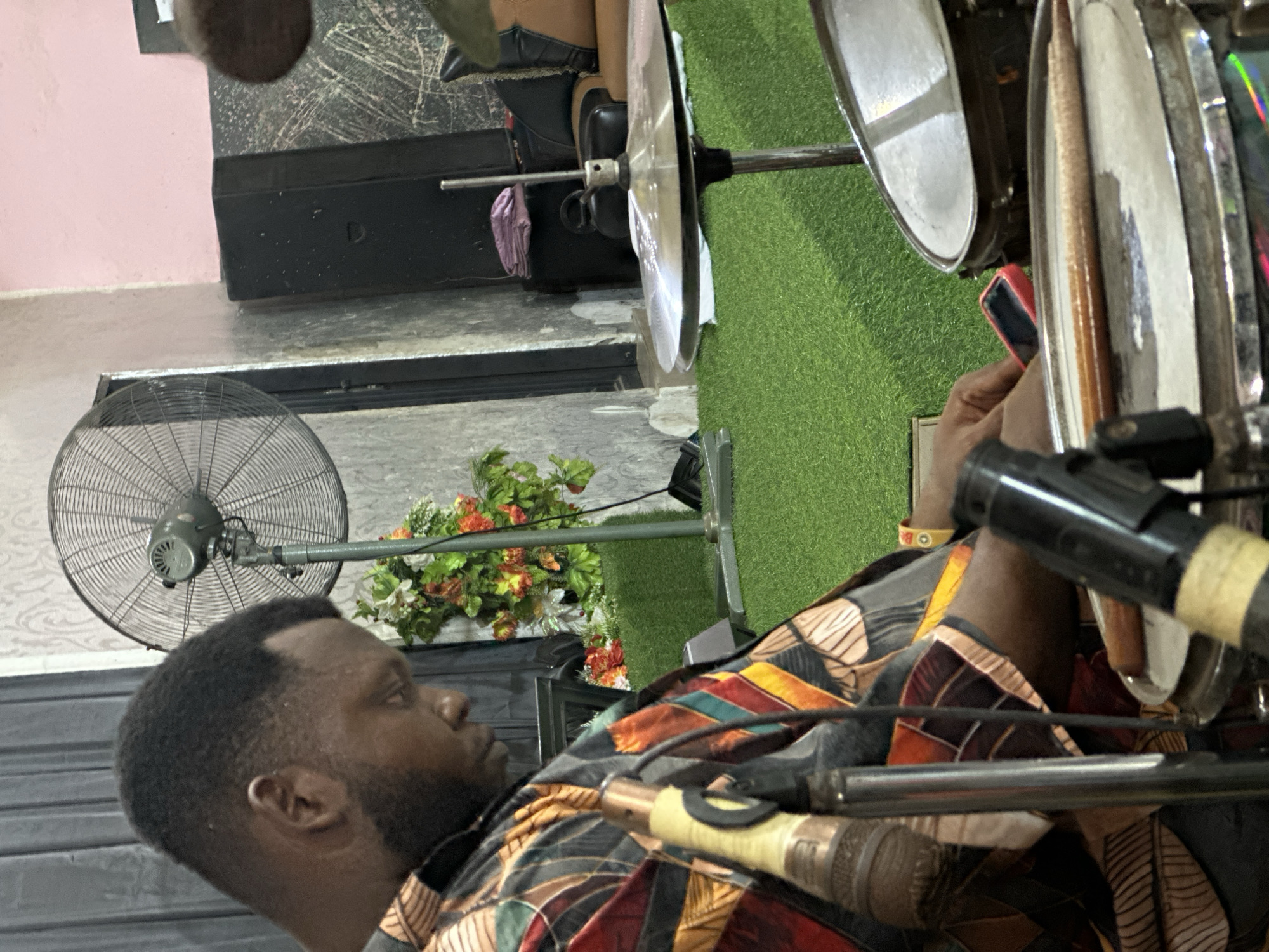 A medium shot of a drummer, presumably in Aba, Nigeria, looking to his left while seated behind a drum set. He is holding a smartphone. To the left of the subject is a green fan with gray metal housing. Behind the fan is a floral arrangement on top of a black amp or speaker. Green turf is seen behind and to the right of the subject. The drummer is wearing a patterned dashiki.