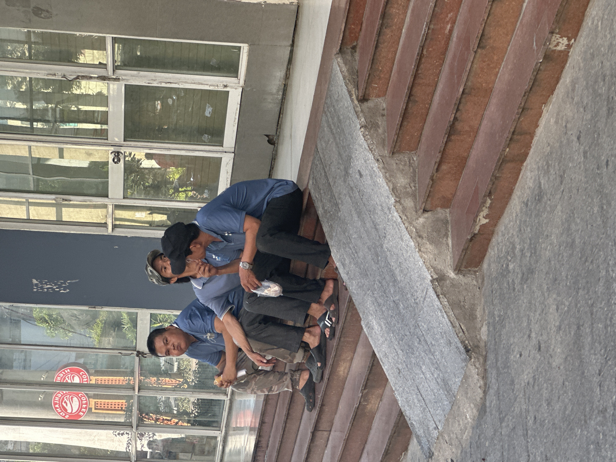 Three men are seated on a set of brick steps outside of a building with glass windows. The men are dressed in matching blue shirts, and two are wearing black pants. One man is wearing camouflage pants. The men are eating. The location is likely Bac Nha Trang Ward, Vietnam.
