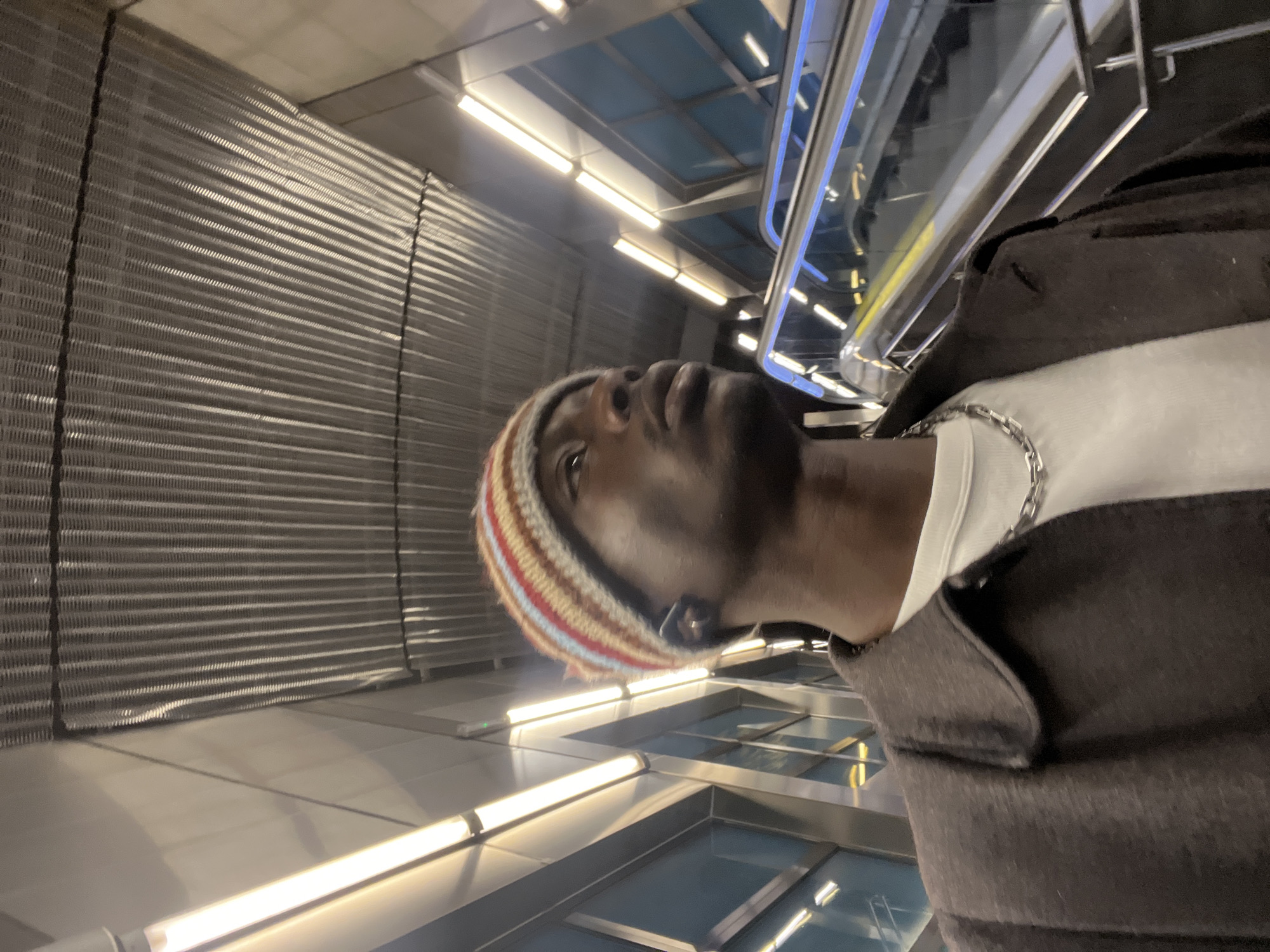 A medium, vertically oriented shot shows a person with dark skin wearing a striped knit hat, a white t-shirt with a silver necklace, and a dark coat. The person is looking upwards. The background includes fluorescent lights, a metal-slatted wall, and an escalator with a transparent barrier. The person is standing in what appears to be a modern subway or train station. I cannot identify the specific location in Barcelona, Spain, without additional information.