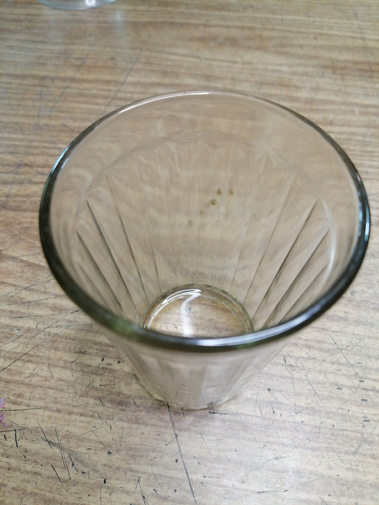 An empty glass, with vertical indentations on its surface, sits on a wooden table. The table is worn, with visible scratches and marks. The glass is positioned at an angle, its rim slightly blurred. The background shows more of the wooden surface. The scene is devoid of any other objects or individuals. There is no indication of location within Rostov-on-Don, Russia, as the image solely focuses on the glass and table.