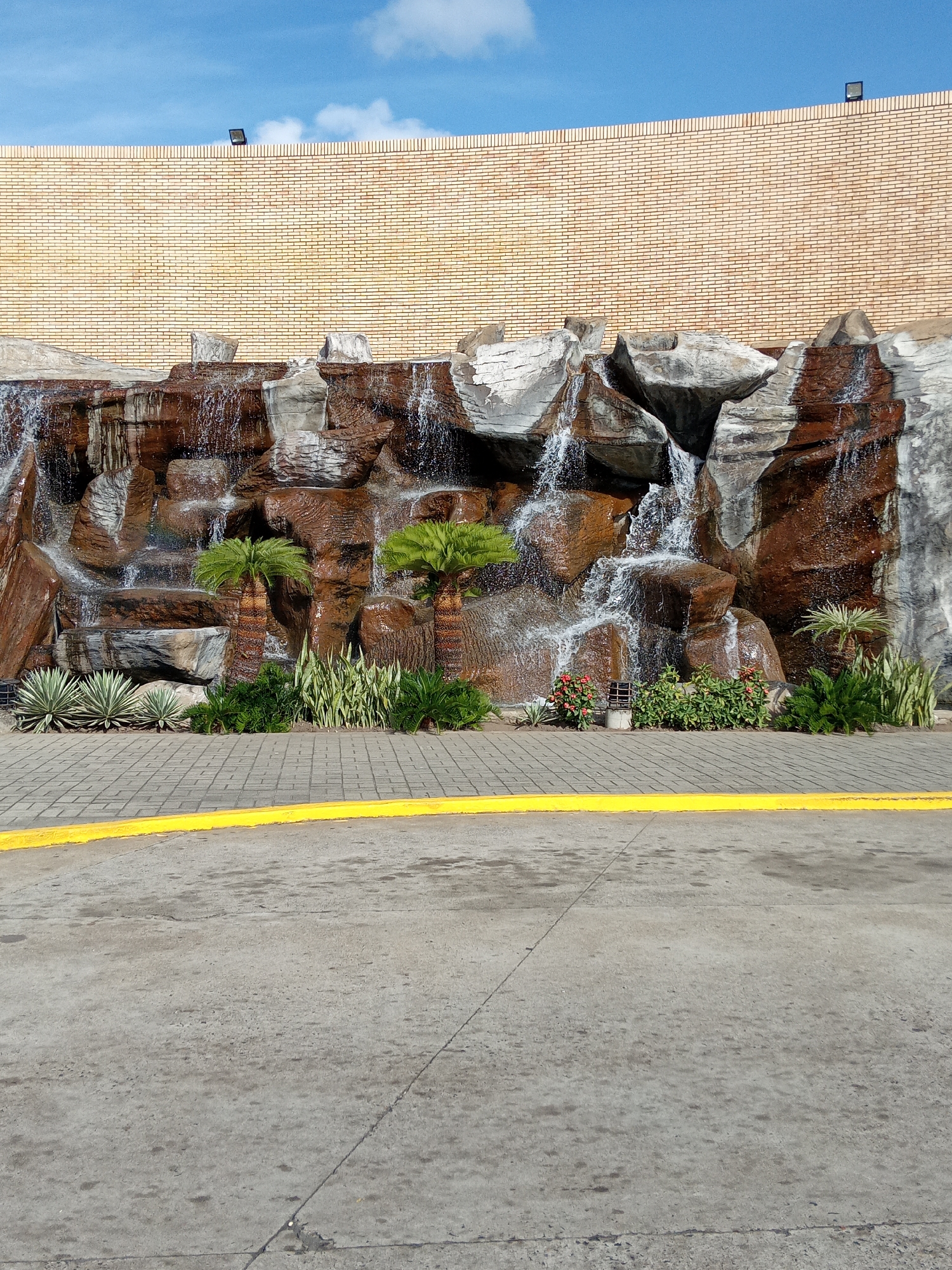A cascading rock waterfall feature is located in Guayana City, Venezuela. The falls are constructed against a high, straight yellow brick wall. Two small, green palm trees are planted at the base. A variety of green plants and succulents are also planted at the base of the falls. Water cascades down the rocks in several places. A yellow line is painted on the grey pavement in front of the falls. The sky above the brick wall is blue with a few white clouds. Two black lighting fixtures are mounted to the top of the brick wall.
