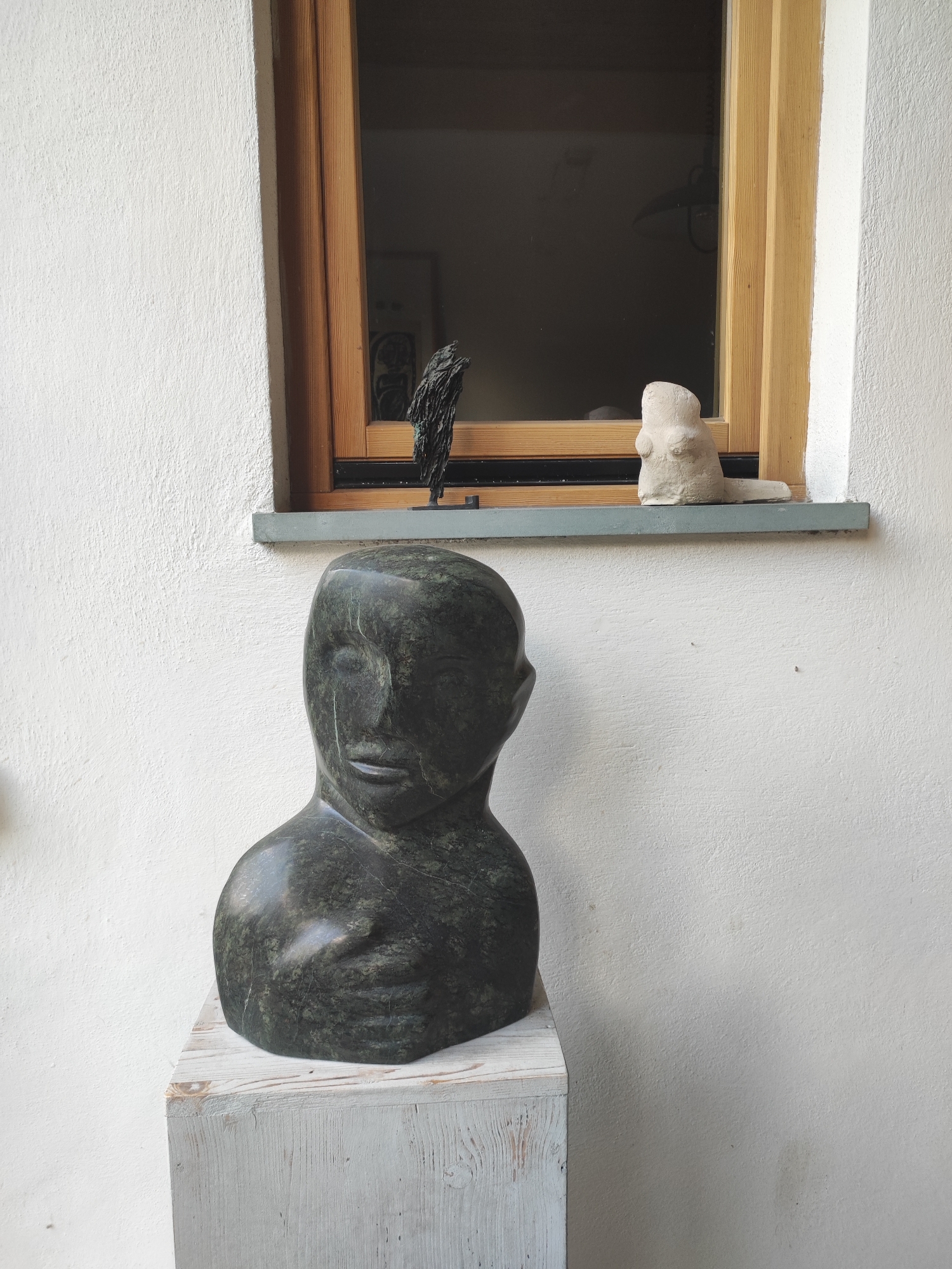 An abstract dark green stone bust of a human head and shoulders stands on a light-colored wooden pedestal against a white stucco wall. To the right, above and behind the bust, is a window with wooden trim, through which a dark interior is visible. On the windowsill below the window, there are two small sculptures: a dark, spiky abstract form and a light-colored, simplified human-like figure. The scene is outdoors, likely in Dresden, Germany, given the architectural style.
