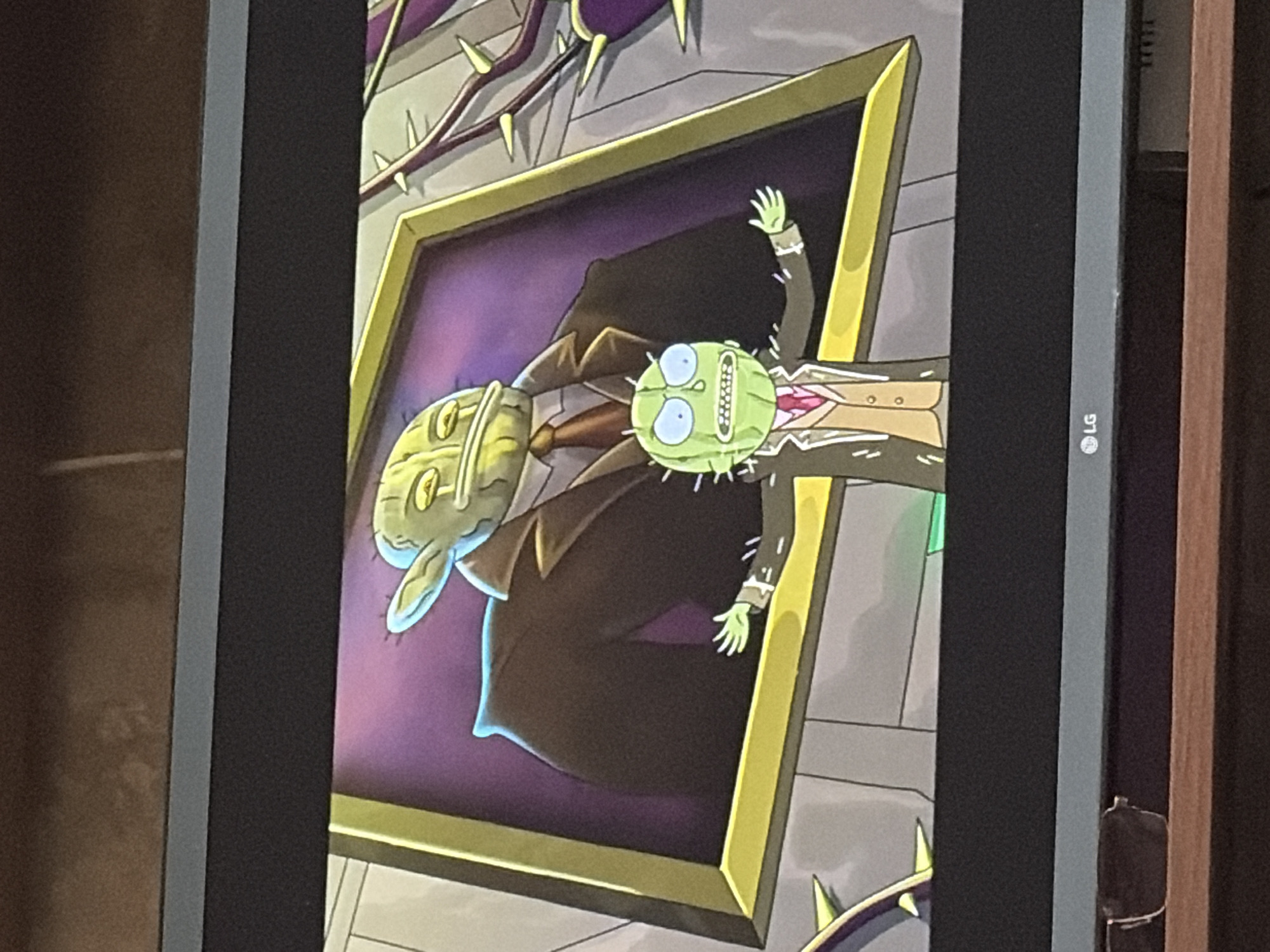 The image shows a television screen displaying a scene from the animated series Rick and Morty. In the foreground, a small, green, cactus-like character wearing a suit and tie stands with its arms outstretched. Behind it, a framed portrait depicts a larger version of the same character, also dressed in a suit, with a purple background. The background of the scene includes elements that appear to be thorny vines and grey walls. The television screen itself is mounted above a wooden surface, and a pair of glasses rests on the surface. The bottom of the television screen shows the LG logo. The scene appears to be an indoor setting, likely a television studio or a stylized depiction within the show's universe.