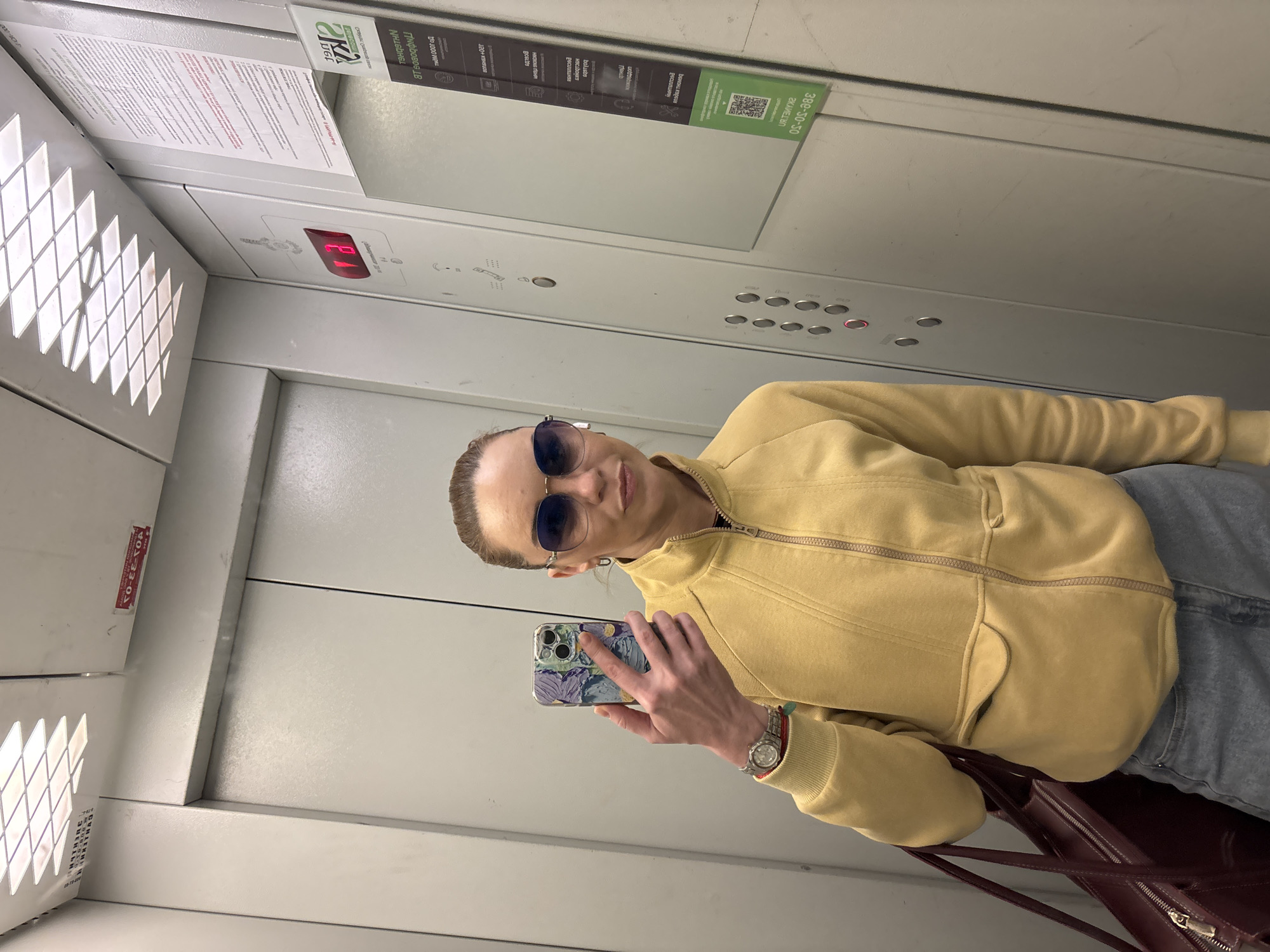 A woman takes a selfie in an elevator. She is wearing a yellow hooded jacket, blue jeans, sunglasses, and a watch. She is holding a smartphone with a colorful case in her right hand. A dark red handbag is visible on her left. The elevator walls are metallic, with visible buttons and a digital display showing "B1." There are various signs on the elevator walls, some in Russian, including what appears to be safety instructions and company information. The overall lighting suggests an indoor setting, likely during the daytime.