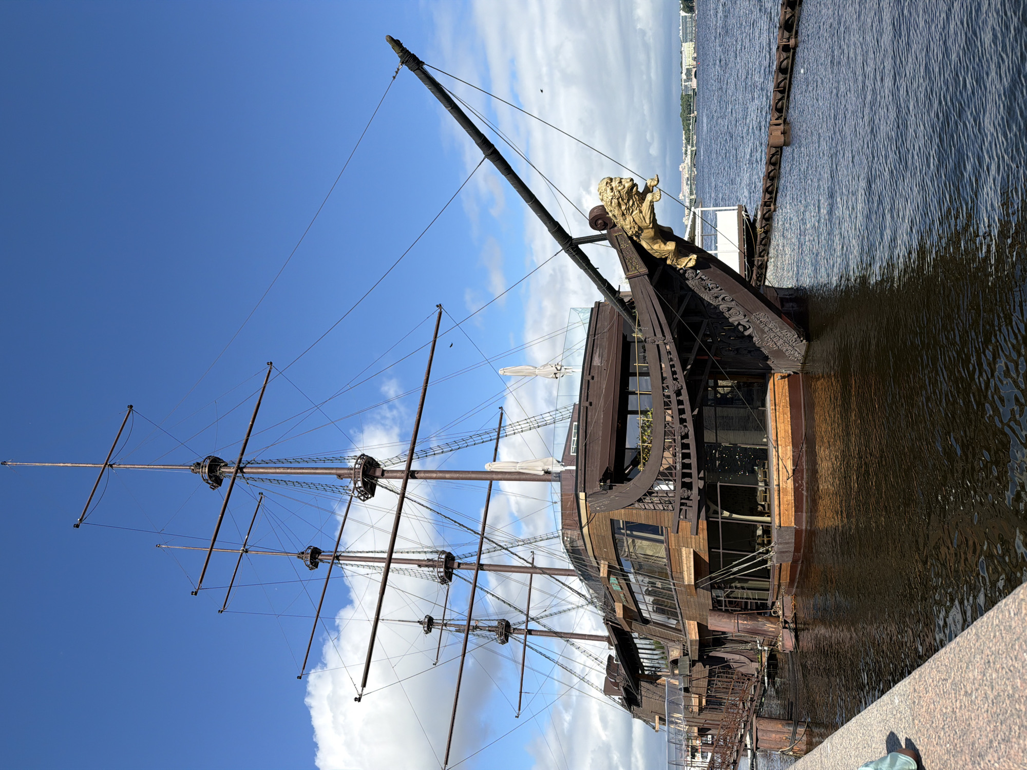 This is a full-frame, eye-level, outdoor shot of a docked, galleon-style restaurant with a wooden exterior, featuring multiple masts and rigging, on a bright, cloudy day. The ship, with a golden lion figurehead, is moored on a body of water, likely a river or canal, in Saint Petersburg, Russia. The water is dark and choppy. In the background, across the water, is a city skyline with several buildings and bridges. The ship is connected to a walkway and is adjacent to a granite embankment. There is no visible activity on or around the ship, suggesting it is not currently open for business.