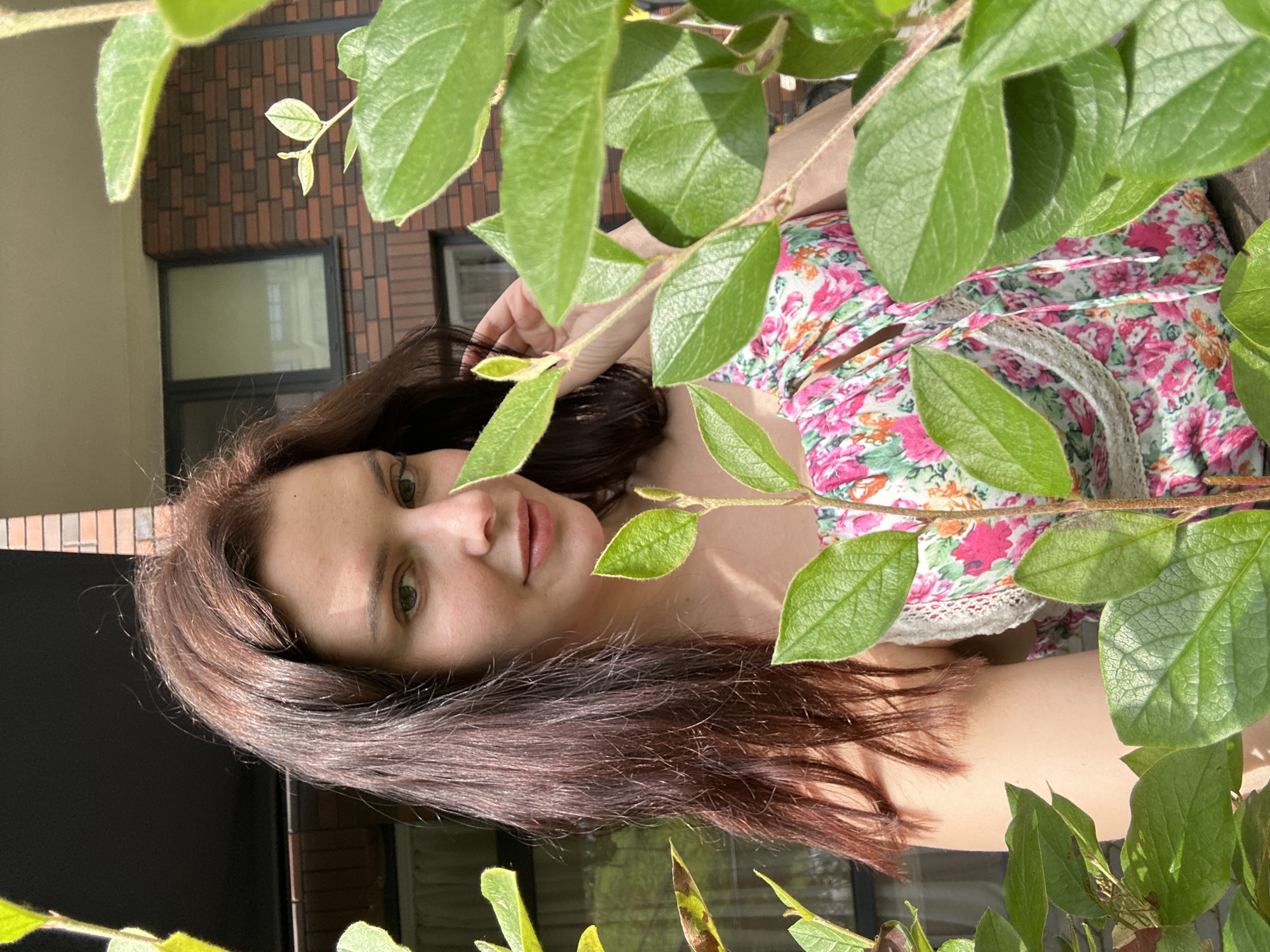 A young woman with dark brown hair is pictured outdoors, partially obscured by lush green foliage. She is wearing a floral-print dress with pink and white flowers. Her head is tilted slightly, and she is looking towards the camera with a gentle smile. The background consists of a brick building with windows. The lighting suggests it is a sunny day.
