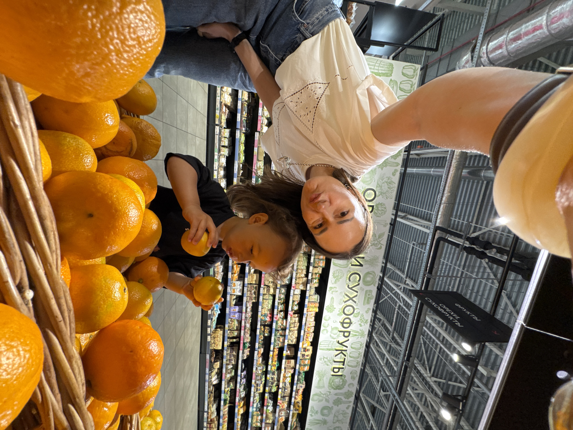 A low-angle, wide shot captures a woman and a young child at a grocery store, likely in Sochi, Russia. The woman, wearing a white embroidered blouse and blue jeans, leans forward, her face partially obscured as she takes the photo. The child, dressed in a black t-shirt, sits in front of a basket overflowing with vibrant oranges, holding two oranges in their hands. Behind them, shelves are stocked with various packaged goods, and a black sign overhead reads "ФРУКТЫ, ОРЕХИ, СУХОФРУКТЫ" (Fruits, Nuts, Dried Fruits). The setting appears to be a modern supermarket with a tiled floor and exposed ceiling infrastructure.