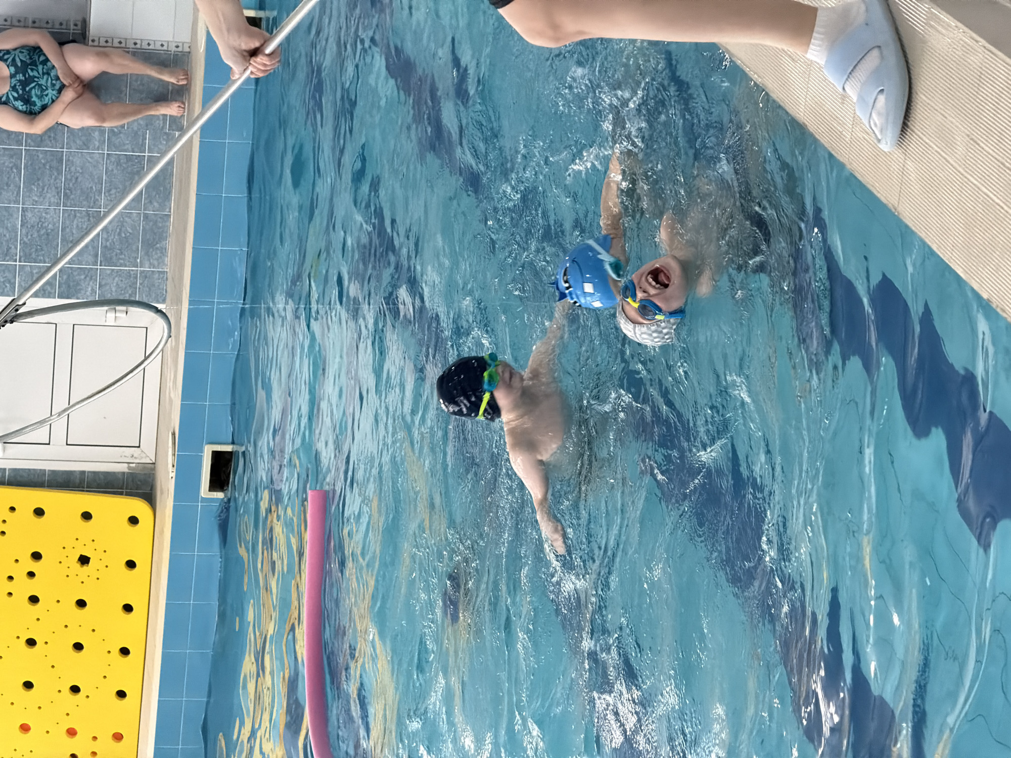 This image captures a vibrant scene at an indoor swimming pool, taken from an overhead perspective and rotated ninety degrees clockwise. Two children, appearing to be boys, are seen in the water, both wearing swim caps and goggles. One child, on the right side of the frame, has a wide-open mouth expressing joy or excitement, wearing a white patterned swim cap and yellow and blue goggles. A blue floatation device or toy is near his head. Another child is further left, partially submerged, wearing a dark swim cap and green and yellow goggles.
The pool itself features clear blue water with visible ripples and reflections, and dark blue lane lines are distinguishable beneath the surface. A pink foam noodle floats near the bottom left corner. Along the edges of the pool, various elements indicate adult presence and pool environment details. An adult's legs and lower torso, clad in a green and blue patterned swimsuit, are visible sitting on the blue tiled floor to the far left. Another adult's hand and arm are seen in the top left, holding a long silver pole that extends towards the pool. In the top right corner, an adult's foot, wearing a light blue slide sandal and a white sock, rests on the textured pool coping.
To the far left, a bright yellow, textured anti-slip mat with numerous holes is visible on the tiled floor. A small white rectangular grate is embedded in the pool wall underwater near the bottom. The setting appears to be a recreational or instructional swimming session, full of active engagement and evident enjoyment from at least one of the children. No visible text is present in the image.