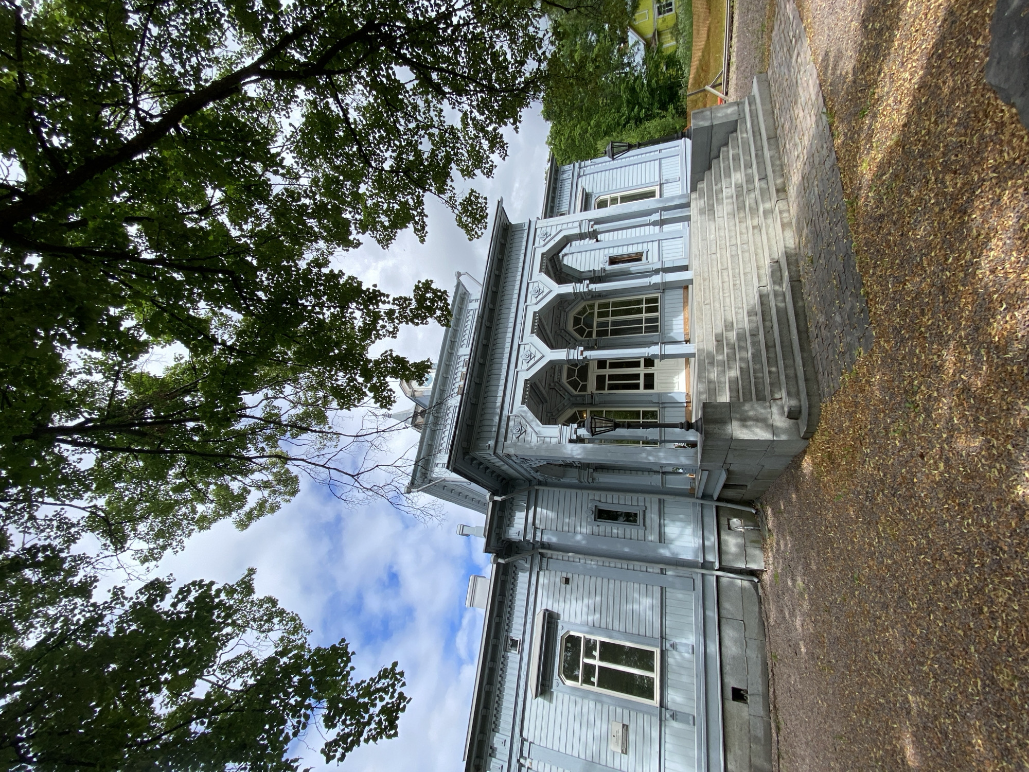 The image depicts a light blue wooden house with a white porch and a grand staircase leading up to its entrance. The house has several windows and ornate architectural details. The surrounding area is covered with gravel and fallen leaves, suggesting an autumn setting. Lush green trees frame the scene, with their branches partially obscuring the sky, which is partly cloudy. In the background, another building with a yellow facade is visible. The house appears to be a historic structure, possibly a museum or a cultural landmark. This scene is likely the Vyborg-based artist Alexander Volkov's house-museum in Sortavala, Russia, known for its distinctive architecture and connection to the artist's life and work.
