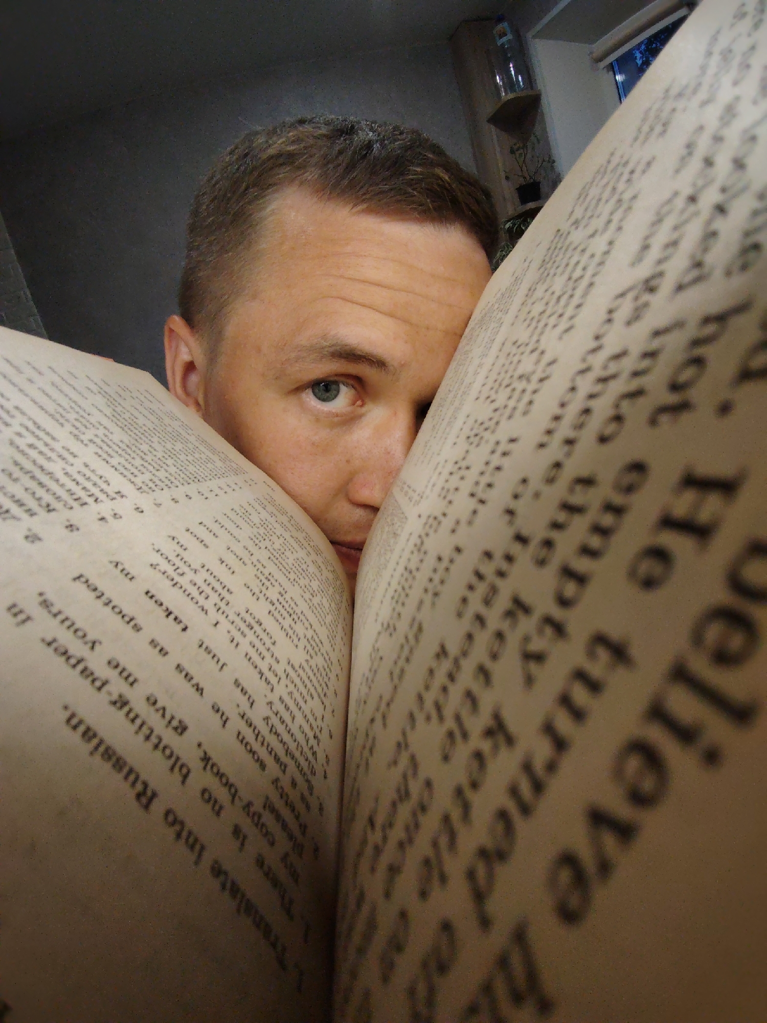A close-up shot depicts a person with short, light brown hair and blue eyes partially obscured by two large open books. The pages of the books are filled with text in a light, sepia-toned paper. Some of the text is in English, including phrases such as "He turned of," "translate into Russian," and "my copy-book." The environment appears to be indoors, with a glimpse of a shelf and window in the background. The focus is on the person's face and the text, creating an intimate and somewhat humorous portrait of someone engrossed in reading or possibly struggling with language learning. There is no indication of the city of Chaikovsky, Russia, or any other geographical location in the provided image.