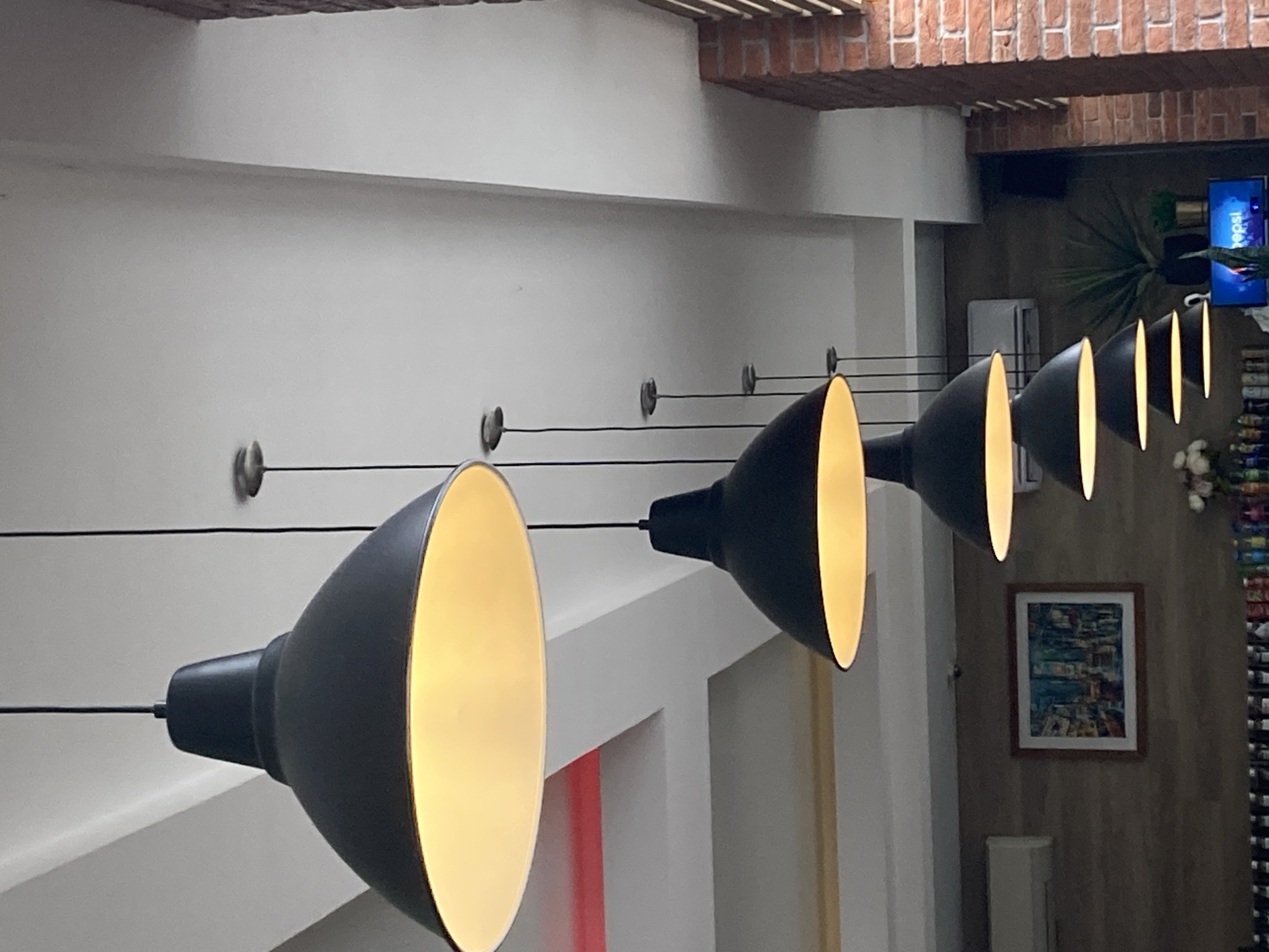 The image shows a row of pendant lights with black shades and gold interiors, hanging from wires against a white wall. The location appears to be an interior space, possibly a restaurant or cafe, with a brick accent on the ceiling. In the background, there is a framed painting on the wall and a television screen displaying a beverage advertisement. The location is possibly in the city of Chaikovsky, Russia.