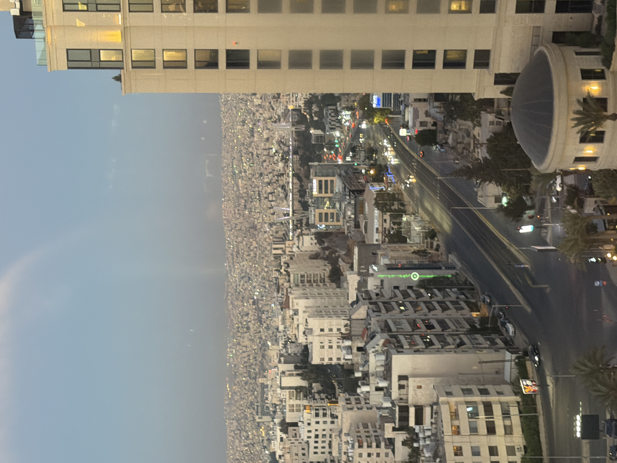 The image captures a panoramic view of Amman, Jordan, at dusk. The foreground is dominated by a modern, light-colored skyscraper with numerous windows, some illuminated. To the left, the cityscape stretches out, a dense collection of buildings in varying shades of white and beige, indicative of Amman's architecture. A highway runs through the city, with cars visible as streaks of light, suggesting ongoing traffic. The sky above is a gradient of pale blue and grey, with a faint hint of a rainbow arching across it. In the distance, the sprawling urban landscape extends to the horizon, dotted with countless lights. A circular, dome-topped structure is visible in the lower right portion of the frame, adding an architectural element to the scene. The overall impression is one of a vibrant, active city transitioning into nighttime.