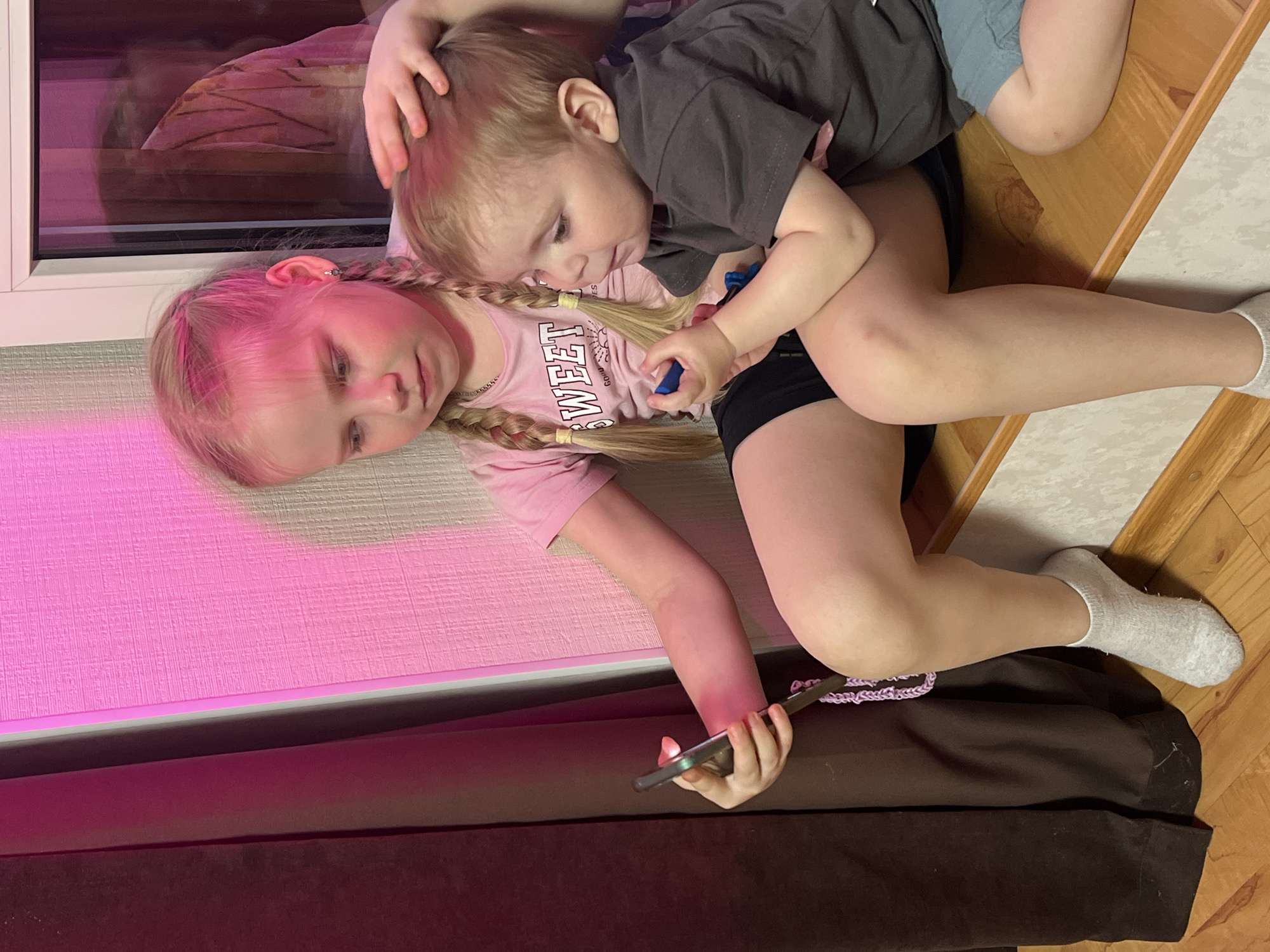 A girl with blonde hair in braids, wearing a pink t-shirt, is lying on a brown and beige floor in Perm, Russia. A small boy with blonde hair, wearing a dark grey t-shirt and blue shorts, is lying on top of her. The boy is holding a blue object in his hand. The girl is holding a dark-colored smartphone. There is a pink wall and a window to the left of the girl. The wooden floor is visible on the right side of the image.