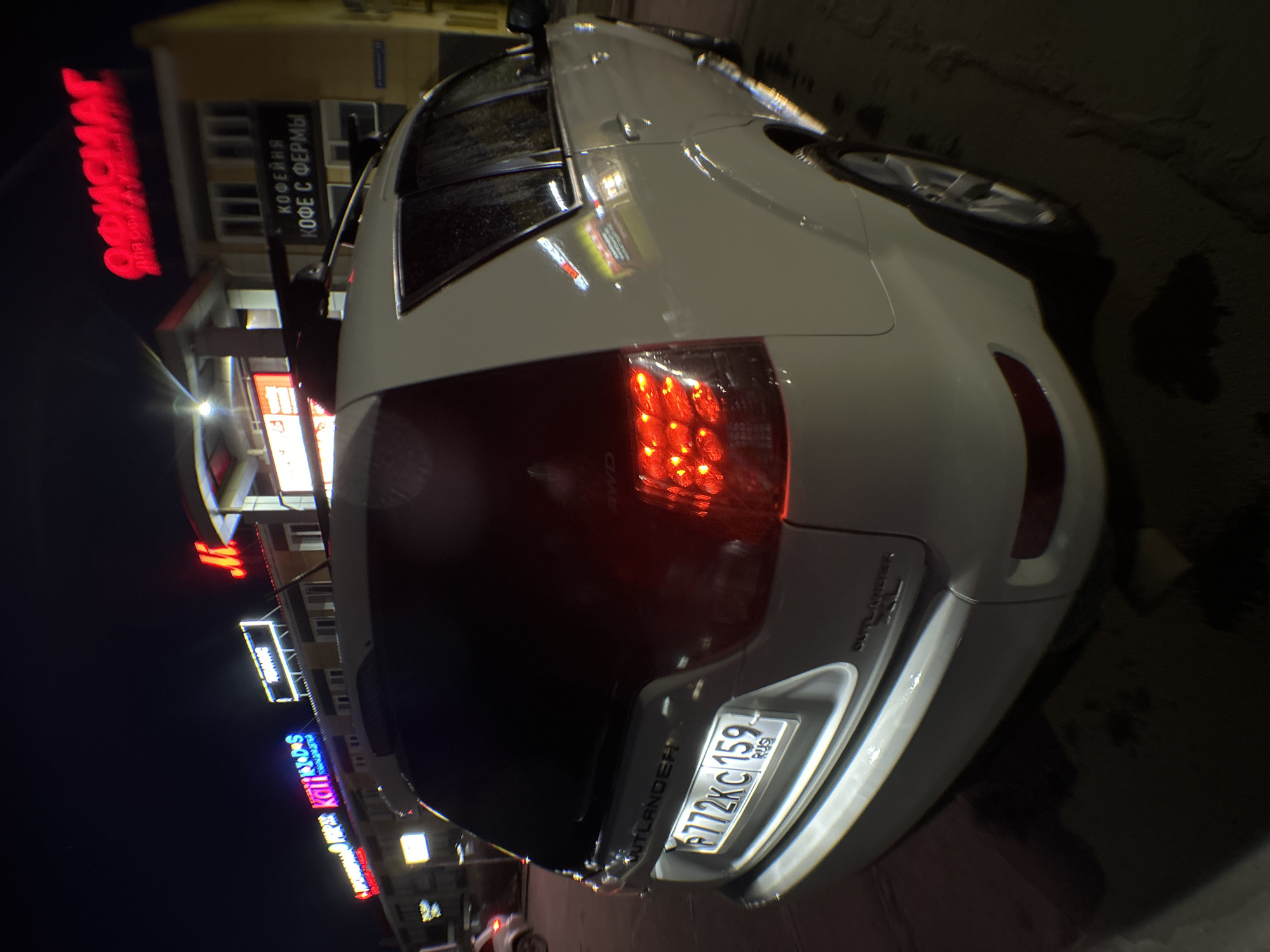 The image depicts the rear of a white Mitsubishi Outlander parked on a street at night. The license plate number is P772KC 159. Streetlights and neon signs are visible in the background, including signs for "KOMPAS," "KOFE S FERMY," and "KALEIDOS." The city is Chaikovsky, Russia, as indicated by the license plate. The environment appears urban. There is no sign of any people or interactions.