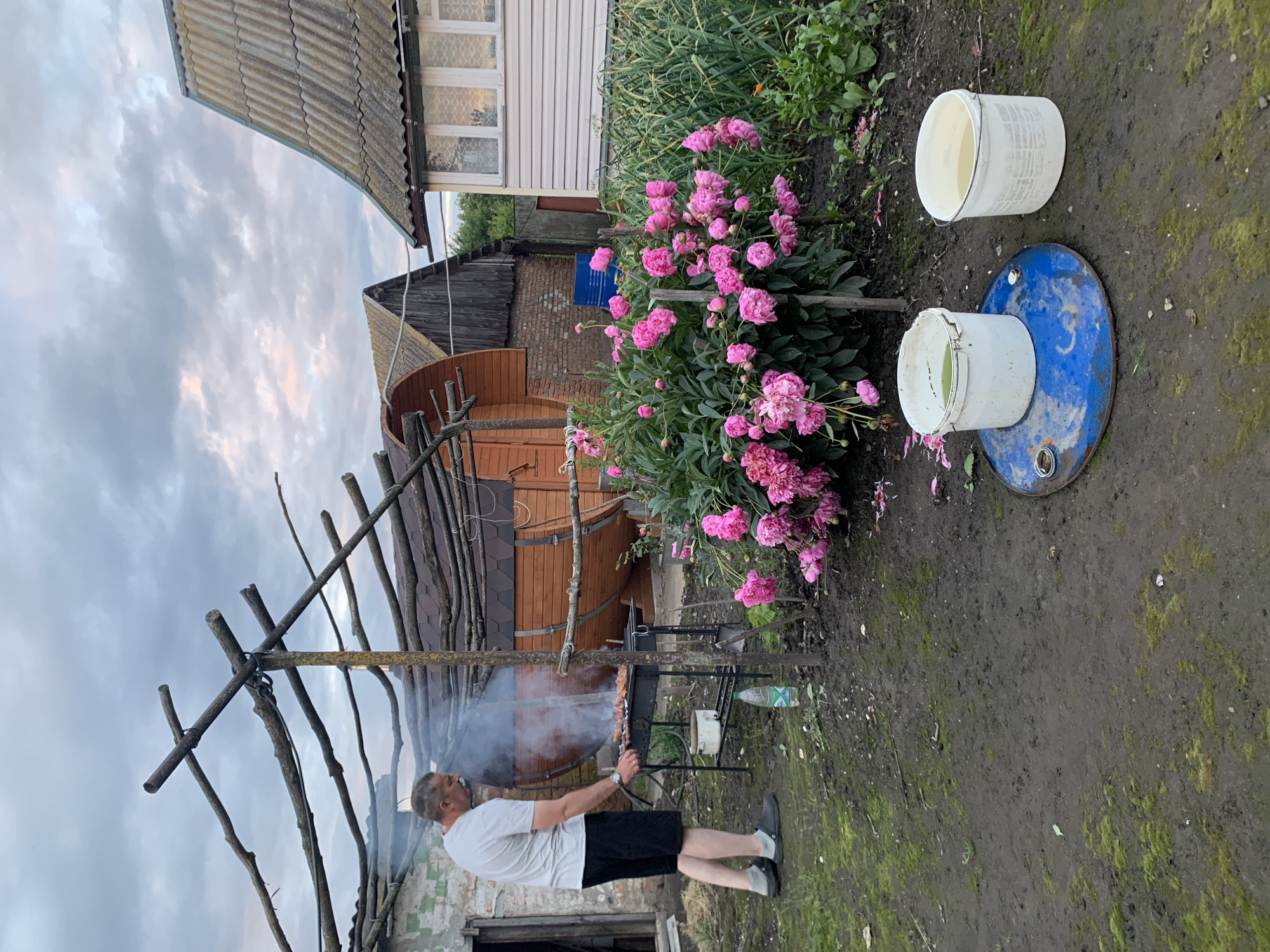 In Блеўчыцкі сельскі Савет, Belarus, a man is grilling meat on a portable barbecue. He is wearing a white t-shirt and black shorts. The barbecue is located on a muddy, overgrown yard. In the background, there are several wooden structures, including a barrel-shaped sauna and a small house with a corrugated roof. A bush with pink peonies is in bloom next to the barbecue. Two white buckets filled with water are on a blue metal barrel in the foreground. The sky is cloudy with hints of pink and orange from the setting sun.