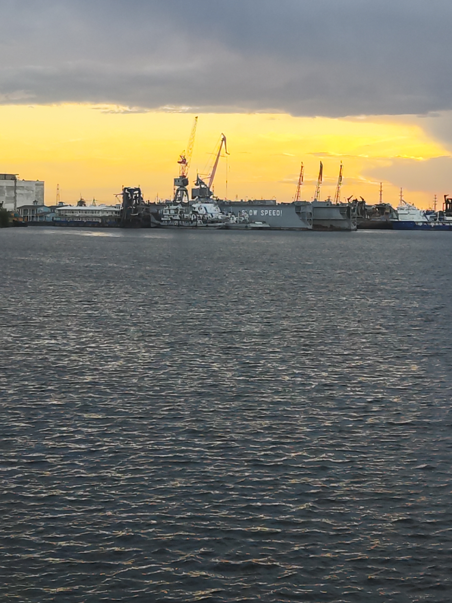 The image shows a waterfront scene at sunset or sunrise, likely in the city of Chaikovsky, Russia. In the middle ground, several large vessels and industrial structures are visible along the shoreline. One large, gray vessel has the phrase "S OW SPEED!" painted on its side. Several cranes are positioned among the ships and industrial buildings. The sky is a mix of dark, cloudy gray and brilliant orange and yellow light. The foreground shows the dark, rippling surface of the water.