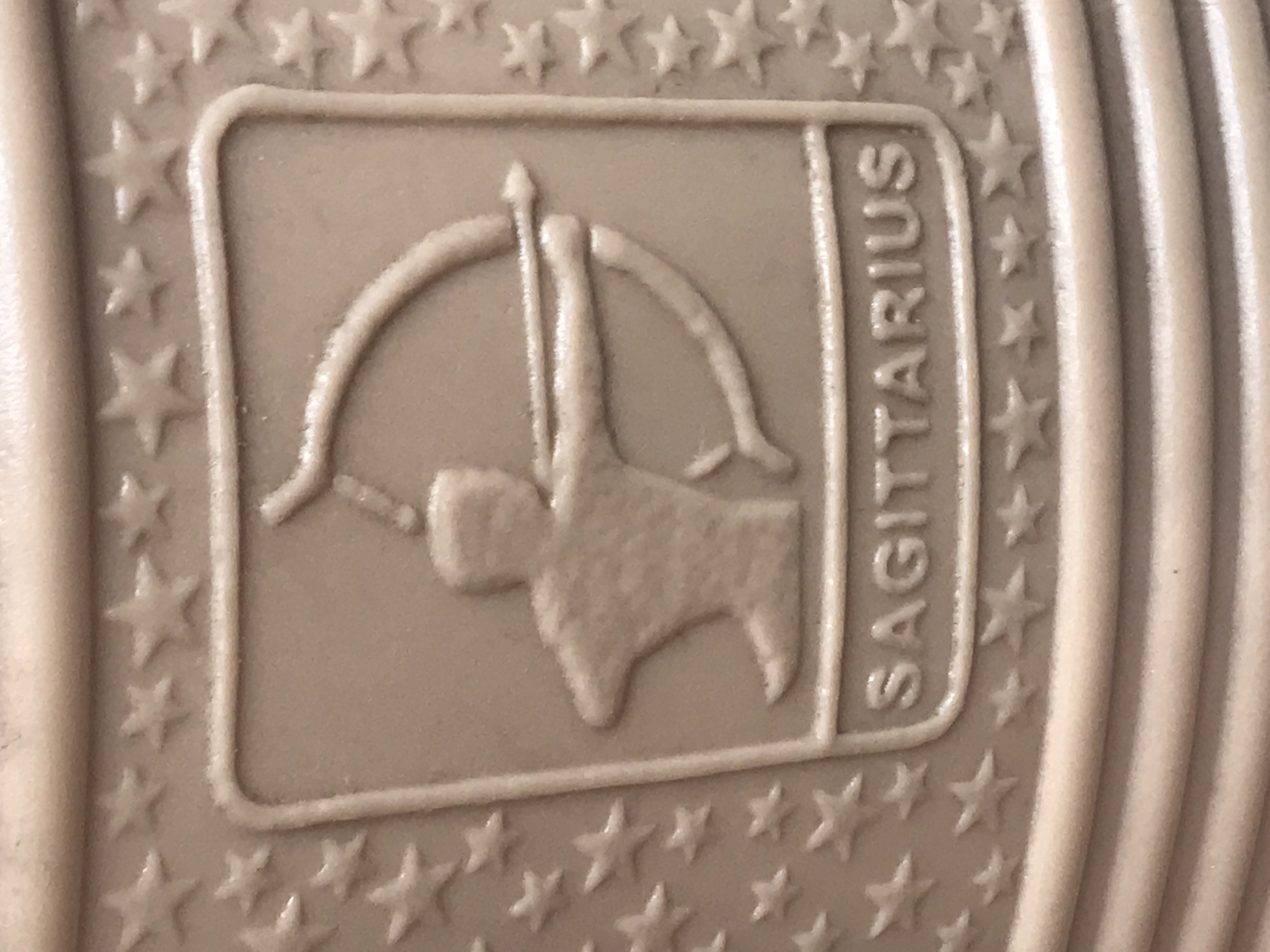The image captures a close-up of an embossed design, likely on the sole of a shoe or a similar molded product, found in Onitsha, Nigeria. The surface is a uniform light brown or beige color, with raised elements creating the design.
The central feature is a rectangular emblem containing a stylized figure representing the zodiac sign Sagittarius. This figure is depicted with a simplified body holding a bow and an arrow drawn. Along the right side of this rectangular emblem, the word "SAGITTARIUS" is embossed vertically in uppercase letters.
Surrounding the central emblem, numerous small, five-pointed stars are embossed in rows, forming a decorative border. To the right of this starred border, the surface transitions into parallel, raised ridges, characteristic of a shoe sole's grip pattern. The material appears to be molded plastic or rubber. There is no specific activity depicted, as it's a static design. The image is well-lit, highlighting the textural details of the embossing.