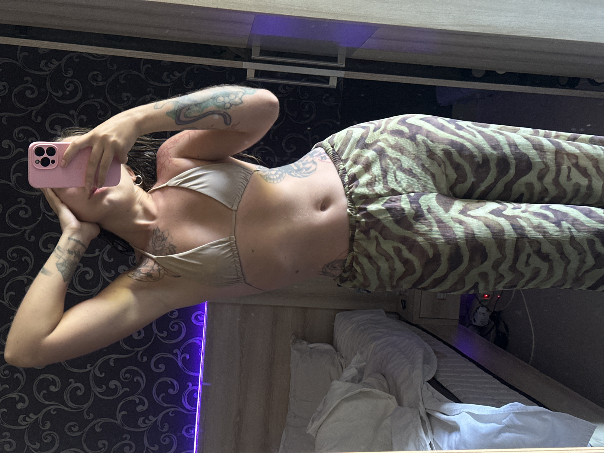 An unidentified female is taking a mirror selfie in a bedroom. She is wearing a light-colored bikini top and patterned pants. Her right arm is raised, and she is holding a pink smartphone to take the picture. The background shows a bed with white linens, a patterned wallpaper, and a glimpse of a closet door. The room is lit by a purple LED light strip above the wallpaper. The location is described as Unknown, Unknown.