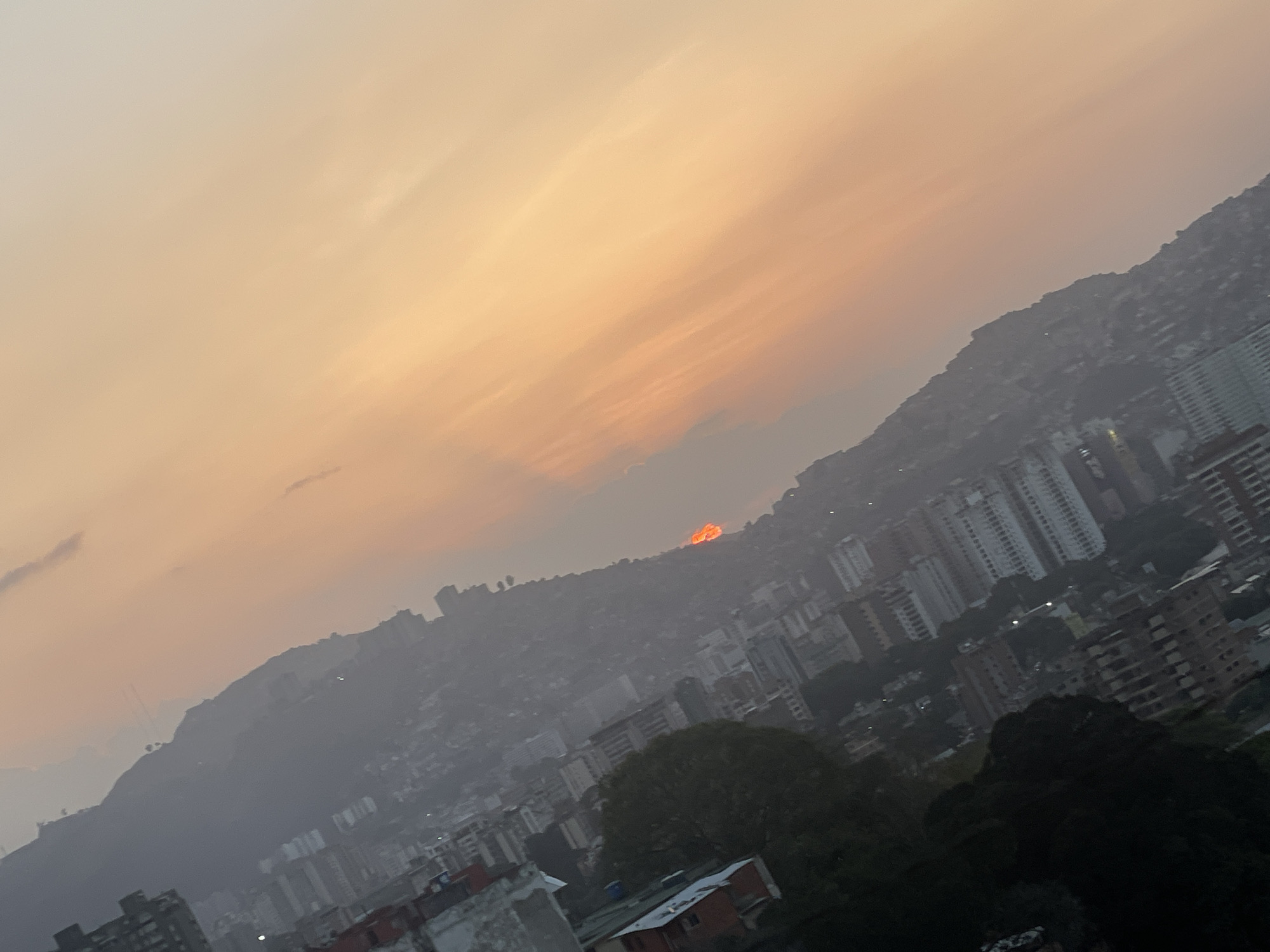 The image depicts a city skyline, likely Caracas, Venezuela, viewed from an elevated position during a hazy sunset or sunrise. The sky is a gradient of orange and pale yellow, suggesting twilight. In the distance, a mountain range, covered in buildings, rises up from the city. The foreground features a dense urban landscape with a mix of residential and commercial buildings. Trees are scattered throughout the cityscape. At the top of the mountain range, the sun is showing around it.