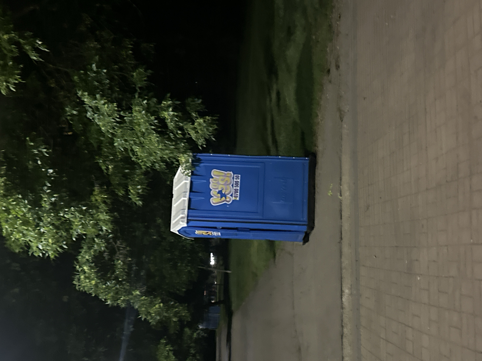 A blue portable toilet with a white roof stands on a paved walkway beside a grassy area with trees. The toilet has a yellow and blue logo with text that reads "0700 500 50". The scene is dimly lit, suggesting it is nighttime or early morning. The walkway is made of reddish-brown paving stones, and a sidewalk is visible to the left of the toilet. The background consists of dark trees and shadows.