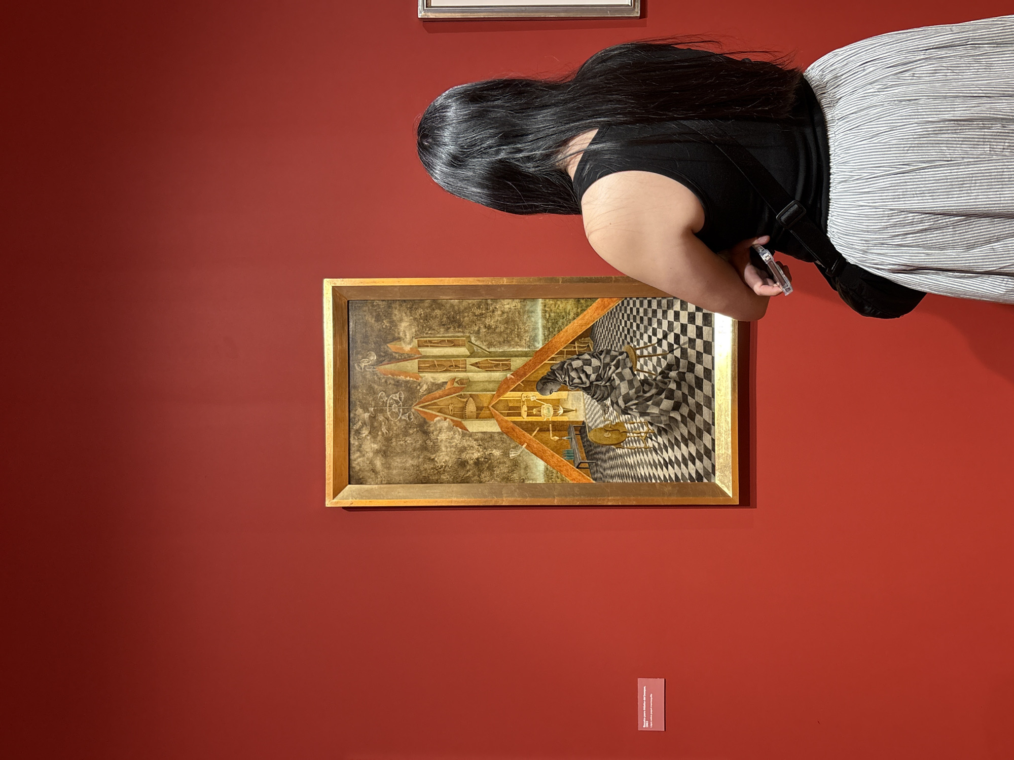 A woman with long, dark hair, wearing a black top and striped pants, stands in front of a painting at a museum in Mexico City, Mexico. She is holding a smartphone and appears to be taking a photograph of the painting. The painting, framed in gold, hangs on a red wall. A small, rectangular sign is affixed to the wall beneath the painting.