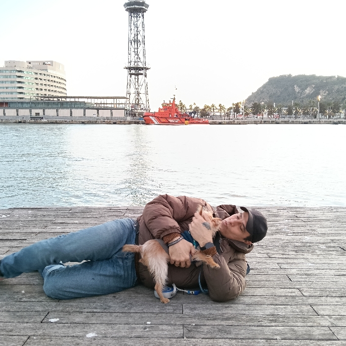 A man in a brown jacket, blue jeans, and a black cap lies on a wooden pier next to the water, holding a small dog. The dog is tan and has a leash attached to its collar. Behind them, there is a body of water with a cityscape in the background. A tall tower and a red boat are also visible. The man appears to be kissing the dog.