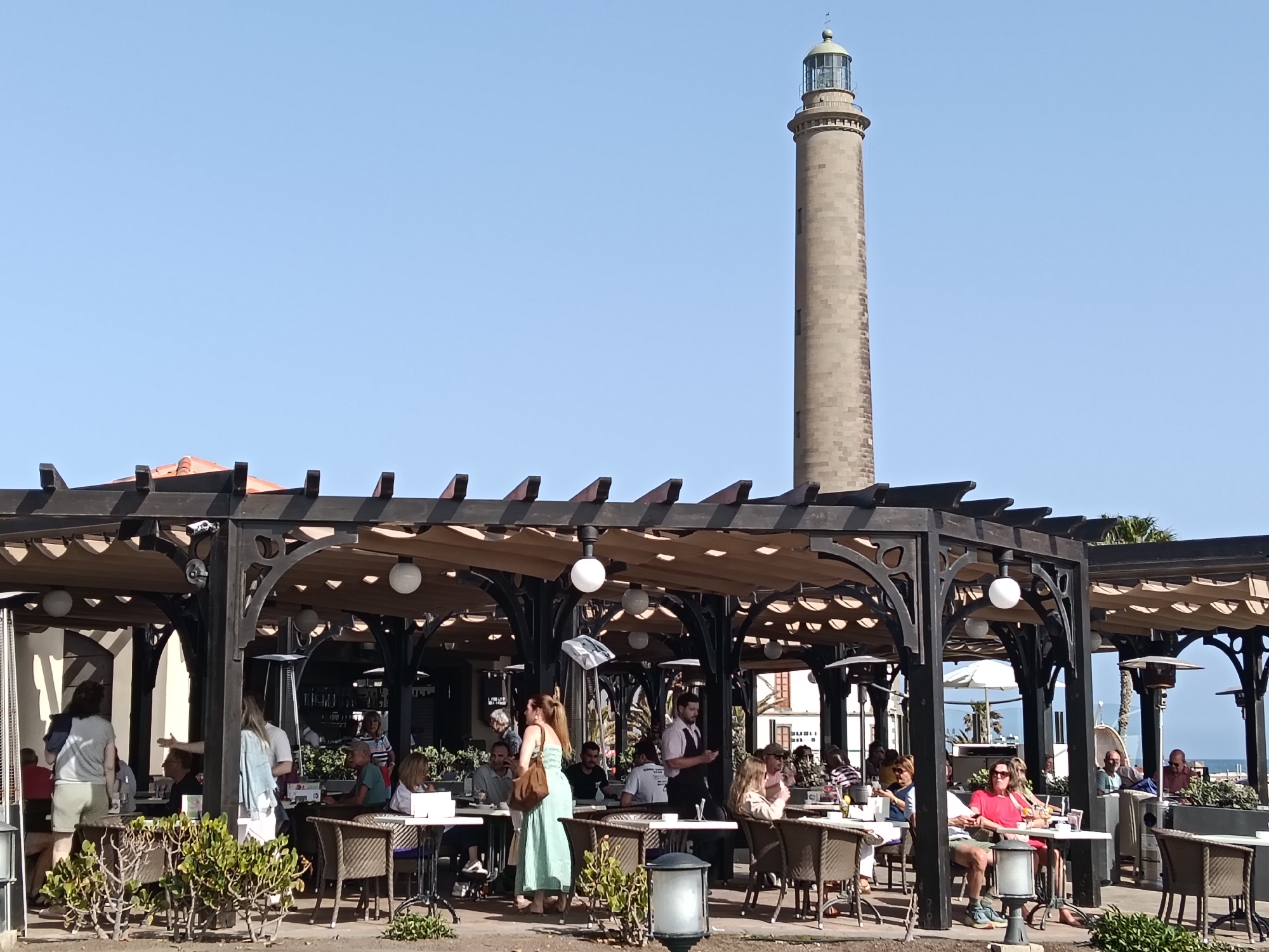 The image shows an outdoor restaurant in San Bartolomé de Tirajana, Spain, during daylight. Patrons are seated at tables under a dark wooden pergola with canvas roofing. The scene is lively, with people eating, drinking, and interacting with a waiter. In the background, the Faro de Maspalomas lighthouse is visible against a clear blue sky. The scene is well-lit.
