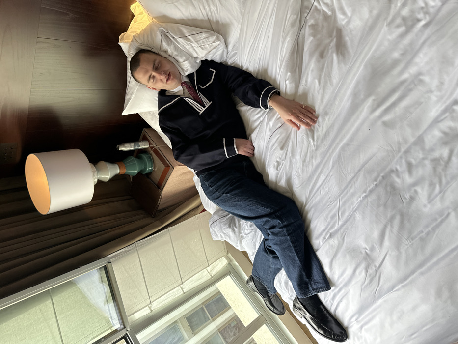 A male individual is observed lying on his back on a large bed in a hotel room in Paris, France. He appears to be asleep or resting, with his eyes closed and mouth slightly ajar. He is dressed in a dark blue long-sleeved top with white trim, a red patterned tie, dark blue jeans, and black loafers. A ring is present on his left ring finger. His head rests on a white pillow, and his left arm is extended onto the white bedsheets. To the immediate left of the bed, a bedside table supports an illuminated table lamp with a white cylindrical shade and a green ceramic base. Adjacent to the lamp are a dark book or folder and a silver spray bottle. A large window on the far left, partially covered by light-colored blinds, displays reflections and glimpses of urban buildings outside. The background wall behind the bed features dark wood paneling.