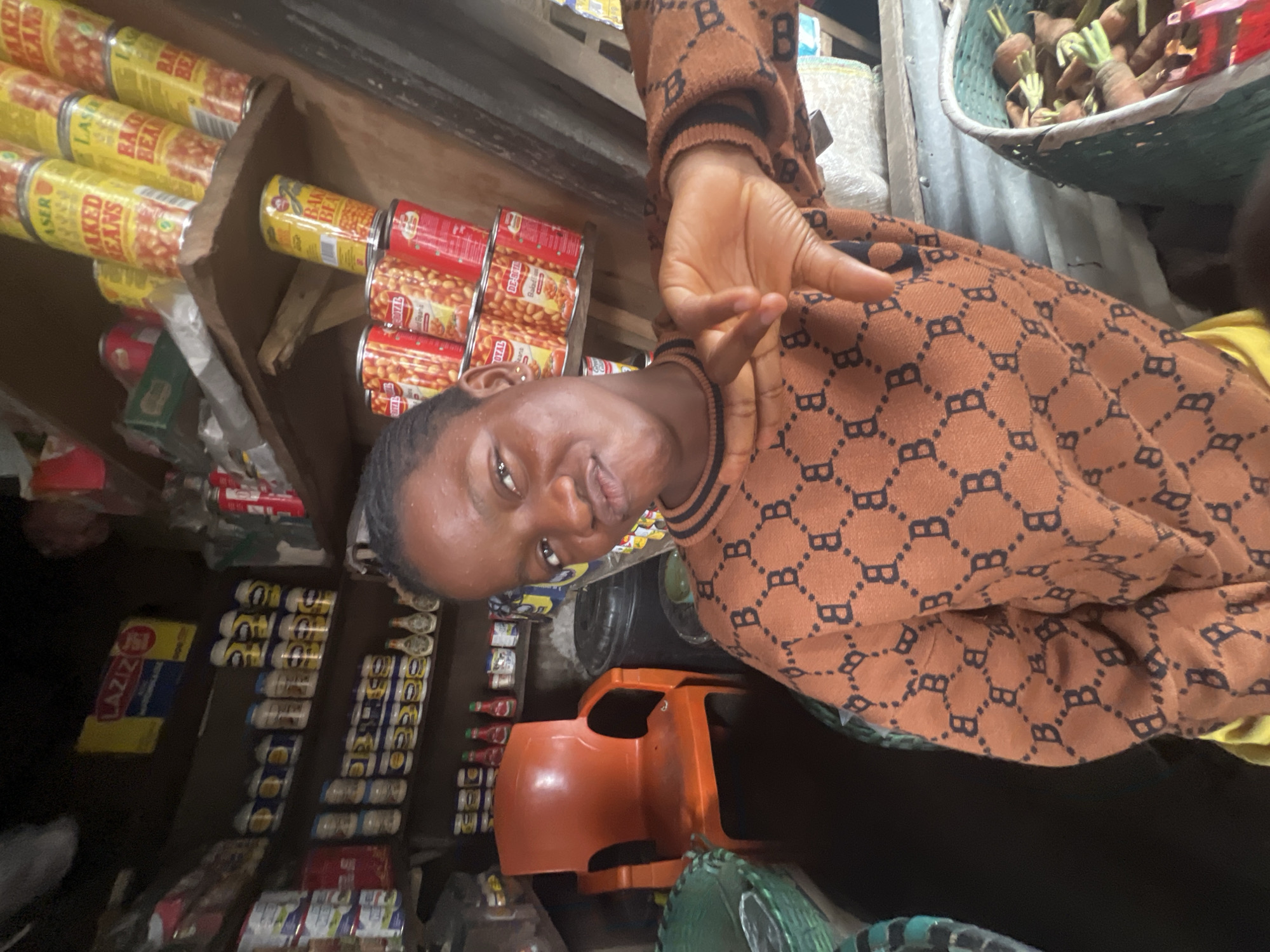 The image depicts a young Black female posing in what appears to be a market stall in Onitsha, Nigeria. She is wearing a brown sweater with a repeating black "B" pattern. Her right hand is raised, with her index finger and thumb forming a "C" shape, and her other fingers extended. The background is filled with shelves stocked with various goods, including canned foods, jars, and bottles. To the left, an orange plastic chair is visible, and to the right, a basket filled with carrots can be seen. The overall environment suggests a bustling marketplace setting.